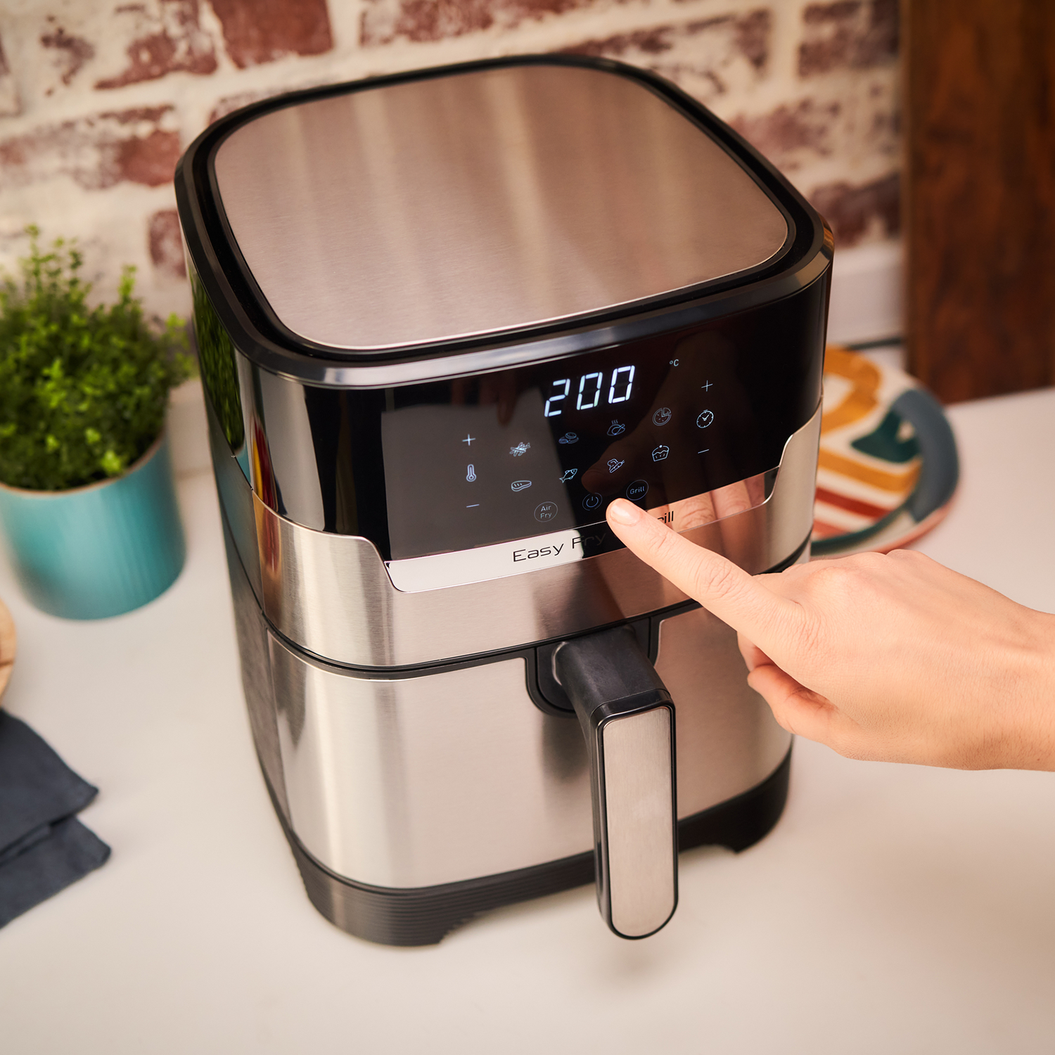 Silver air fryer with a black display, showing 200. Finger presses a button. Background: brick wall.