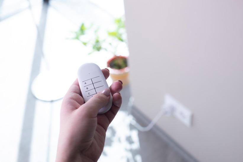 Hand hält eine weiße Fernbedienung mit den Tasten A, B und C. Eine Steckdose ist im Hintergrund zu sehen.