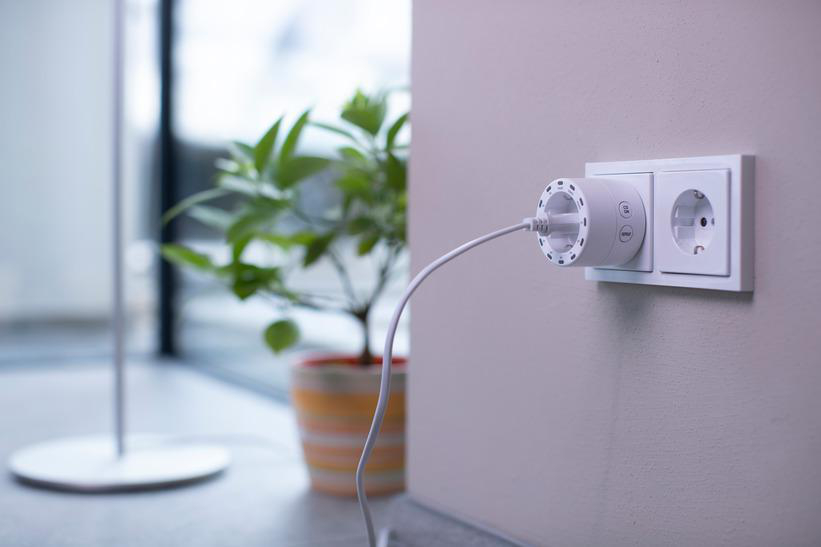 Weißer Smart Plug in einer Steckdose. Eine Pflanze im Topf ist in der Nähe. Wand ist hellrosa.