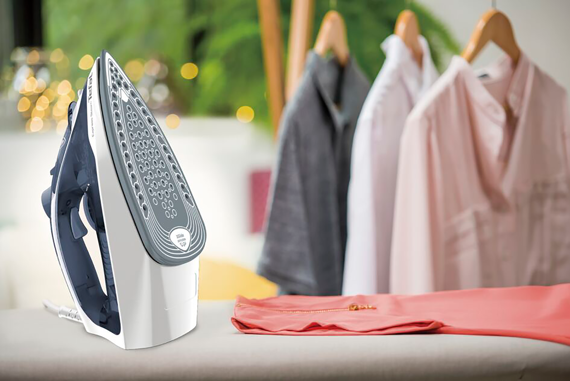 White and gray iron on an ironing board. Clothes hanging in the background.