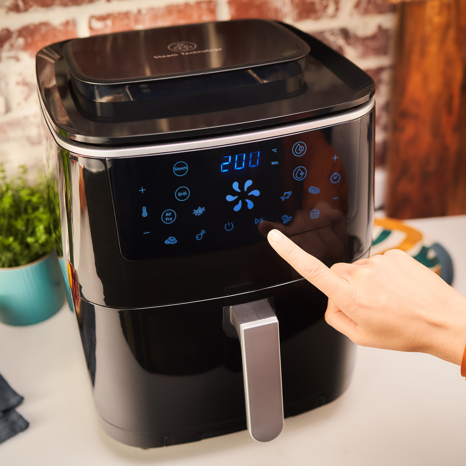 Zwarte airfryer. Hand raakt het display aan, toont '200'. Wit aanrecht, wazige achtergrond.