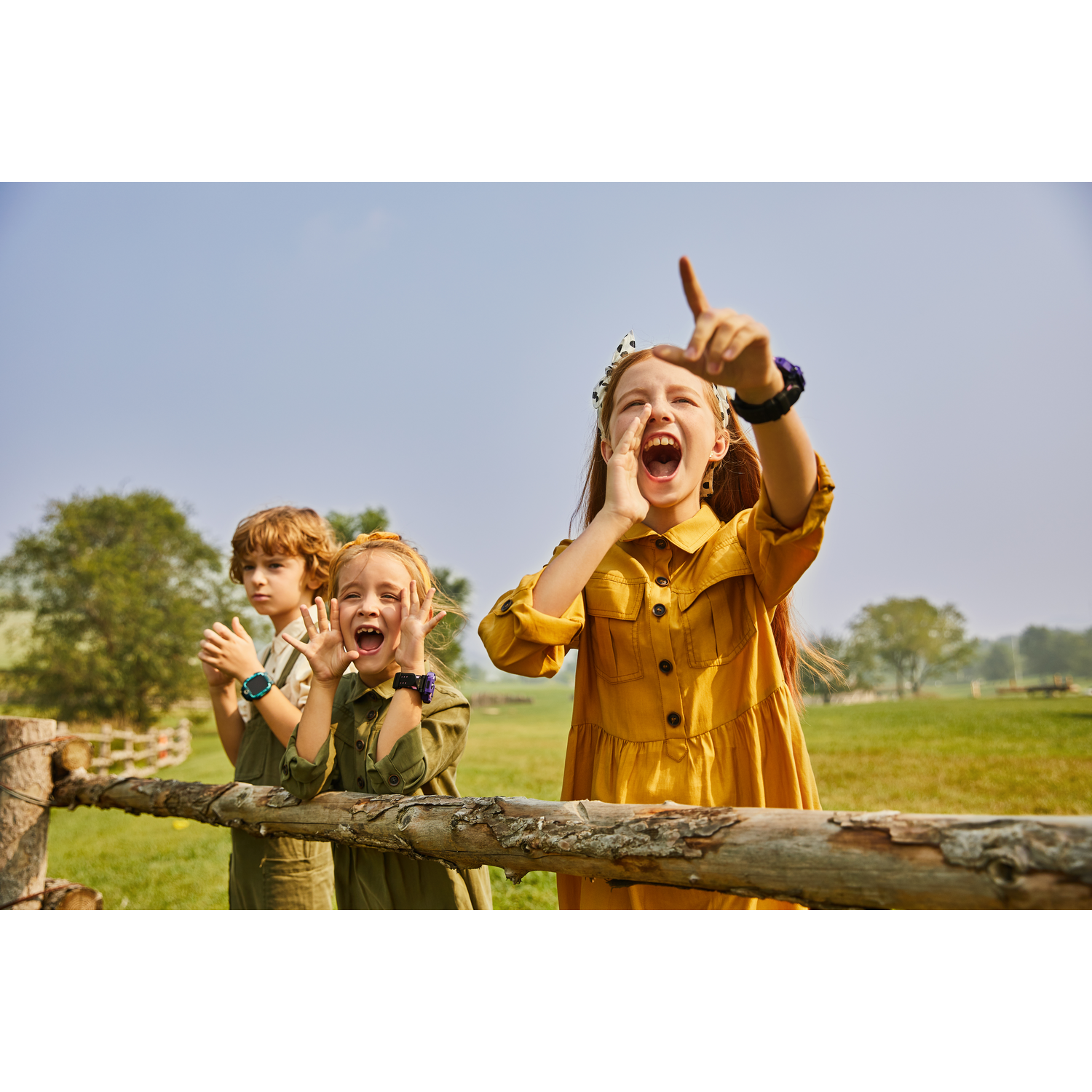 Tres niños en un campo, uno señalando hacia arriba y gritando. Árboles y valla al fondo.