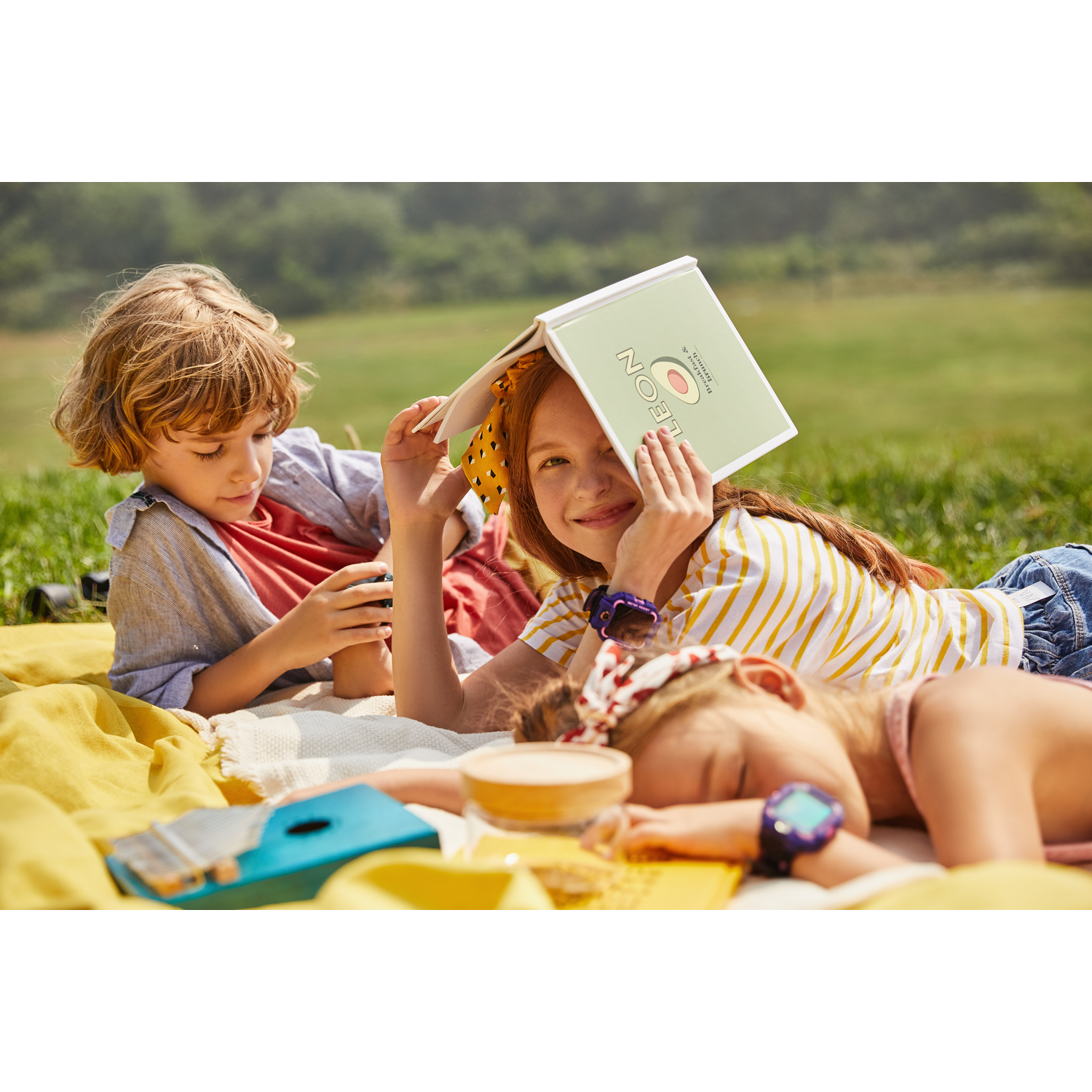 Tres niños en una manta, leyendo y jugando. Uno se cubre la cara con un libro.