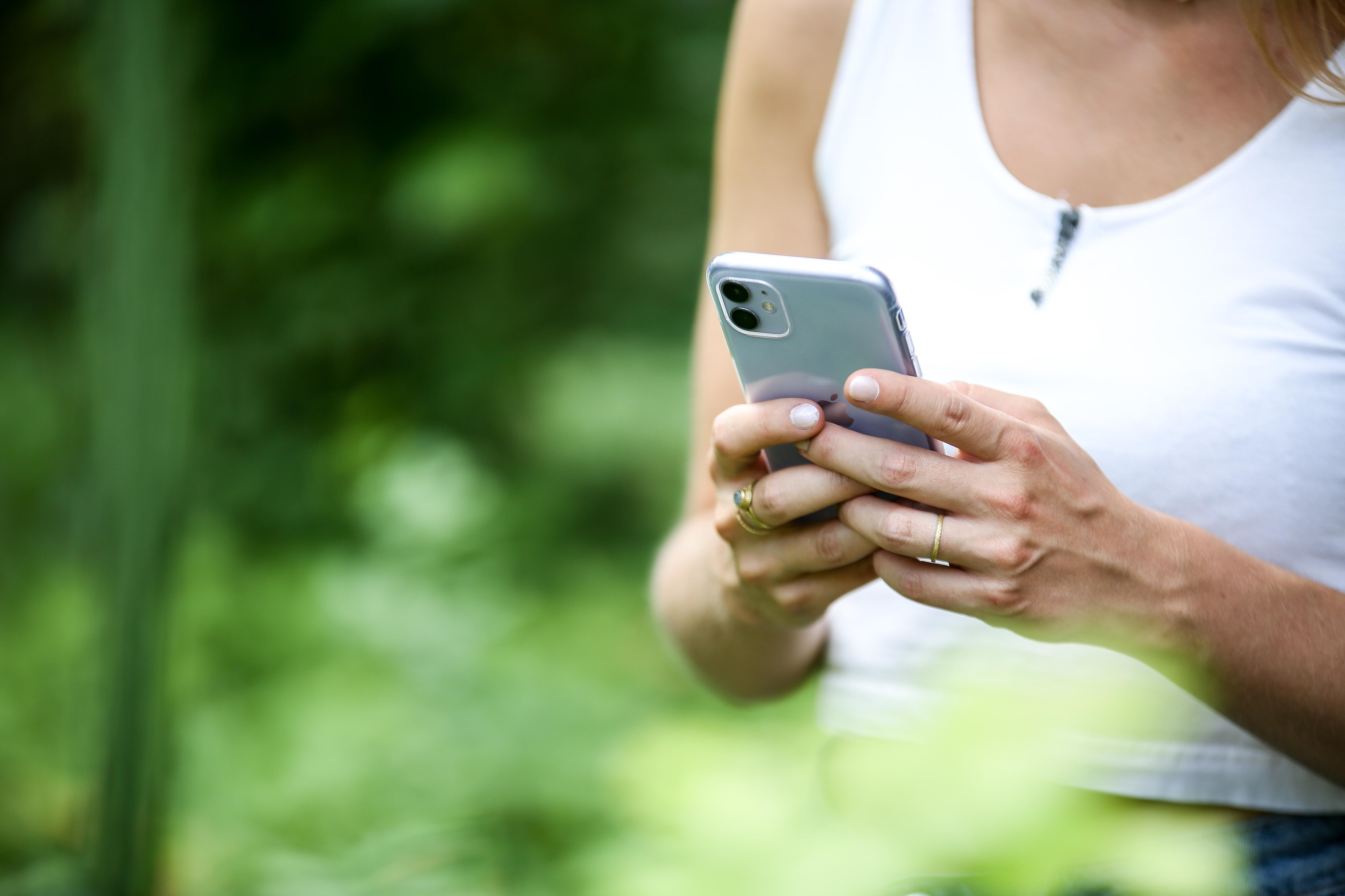Frau hält ein Telefon in einem grünen Garten, trägt ein weißes Oberteil, verschwommener Hintergrund.