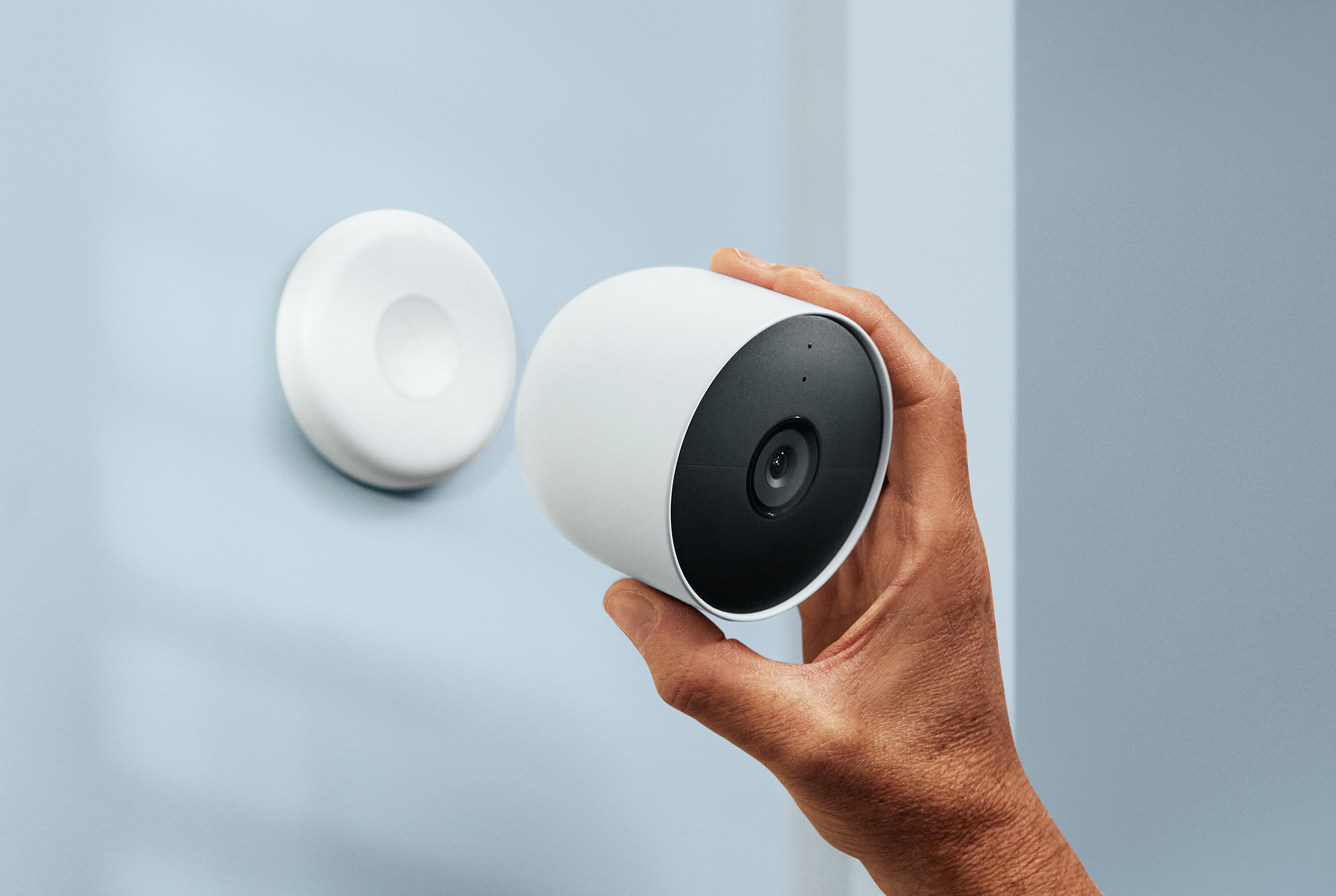 A hand holding a white security camera next to a mounting base on a blue wall, showing installation.