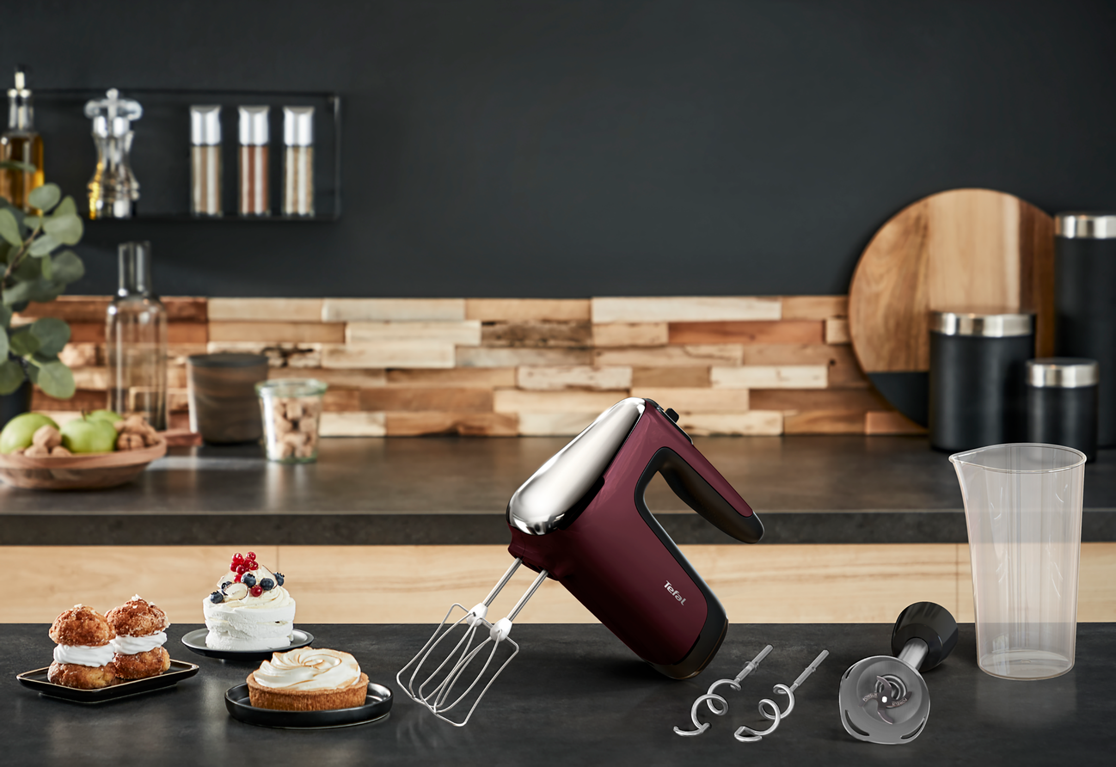 Burgundy hand mixer on a countertop with cakes and ingredients. Dark wood background.
