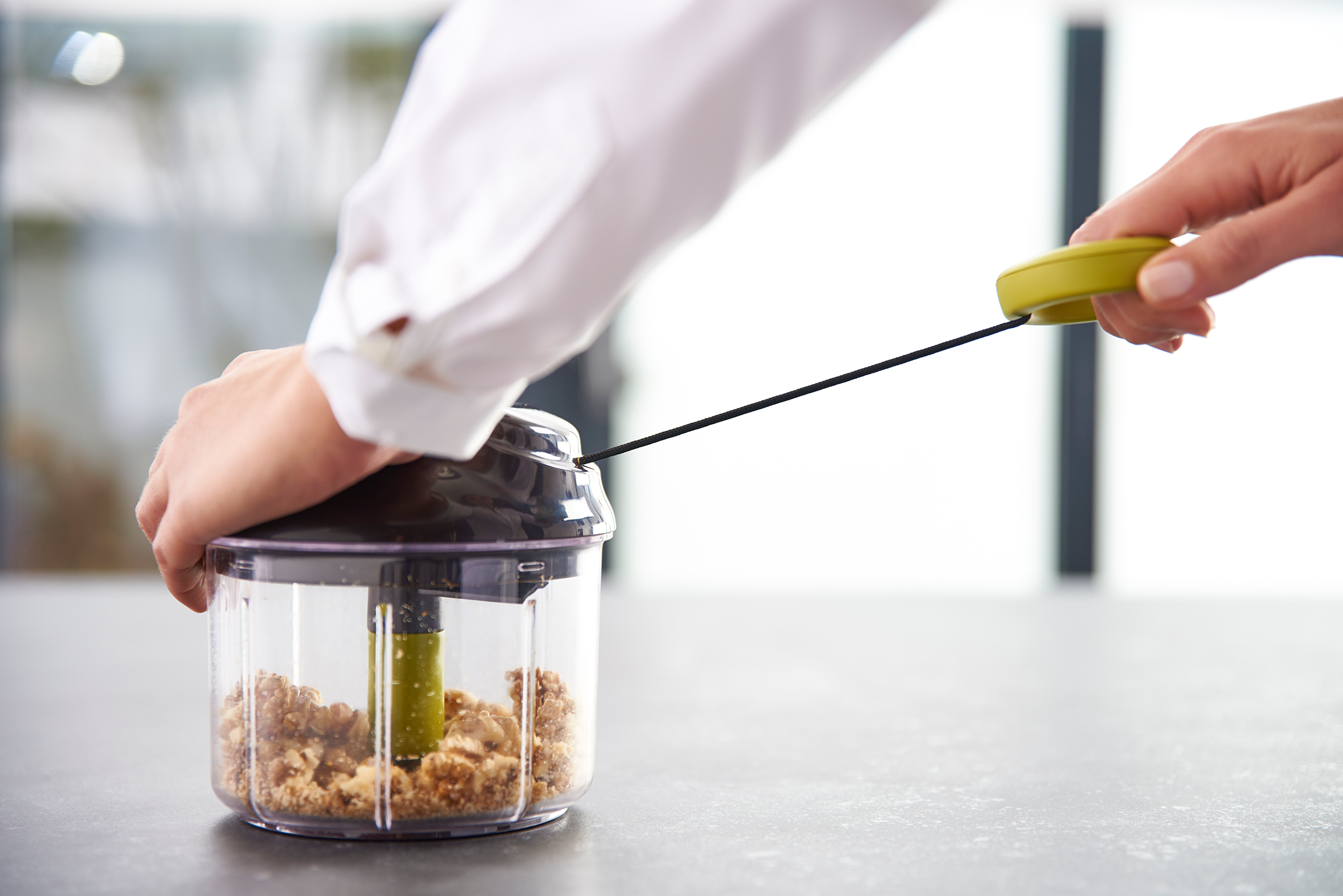 A person using a pull-cord chopper. The chopper is transparent and contains chopped ingredients.