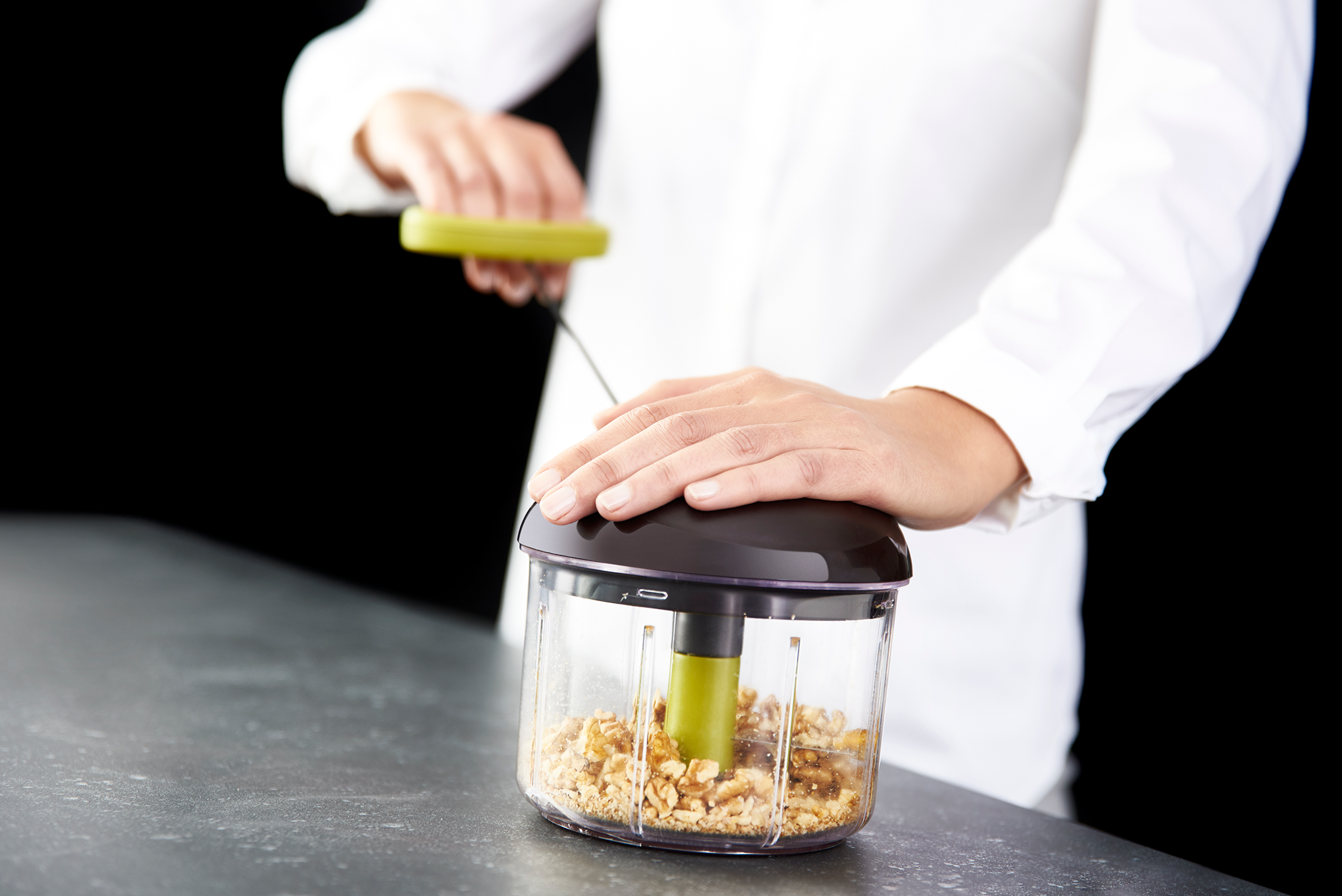 A person using a green-handled food chopper with walnuts inside against a black background.