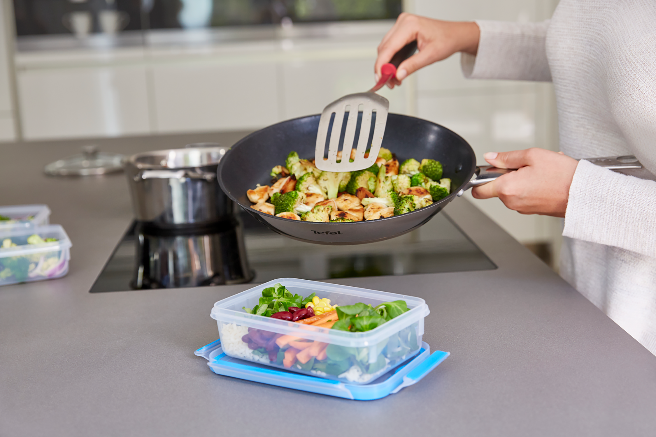 Person cooking. A pan with food, a container with salad on the counter. Kitchen in background.