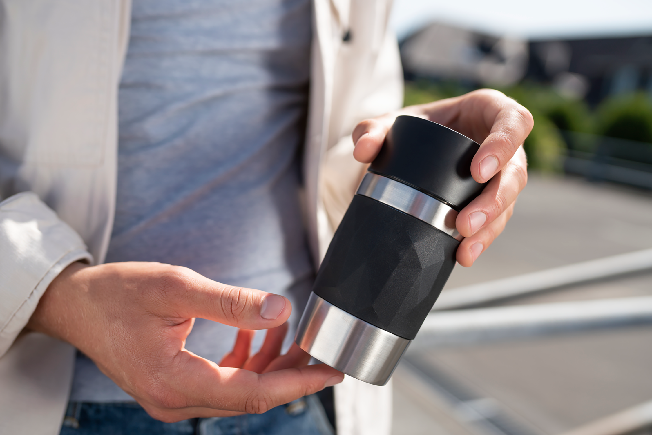 A person holding a black and silver travel mug outdoors on a sunny day.