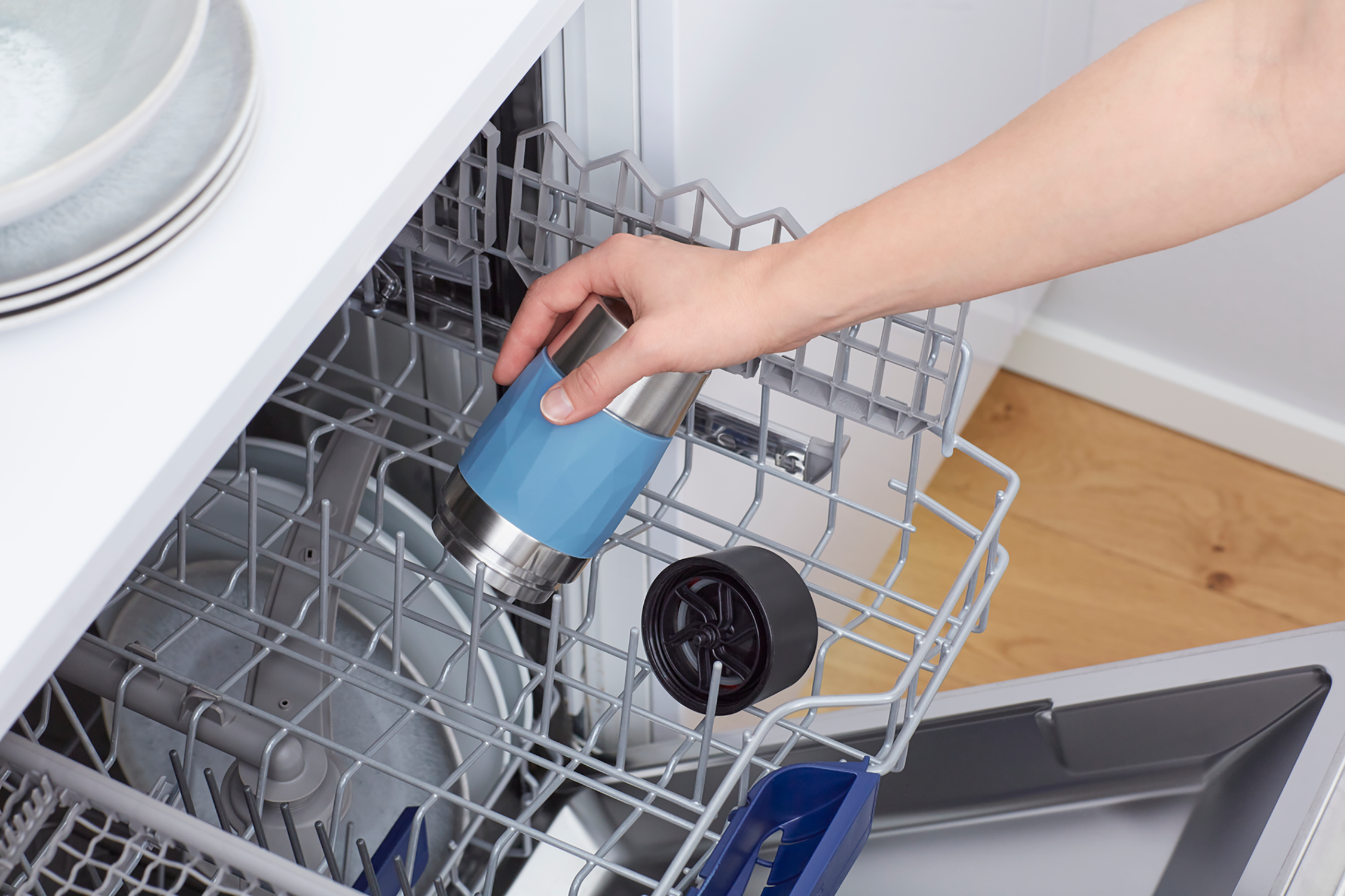 A hand placing a blue and silver tumbler into a dishwasher rack.