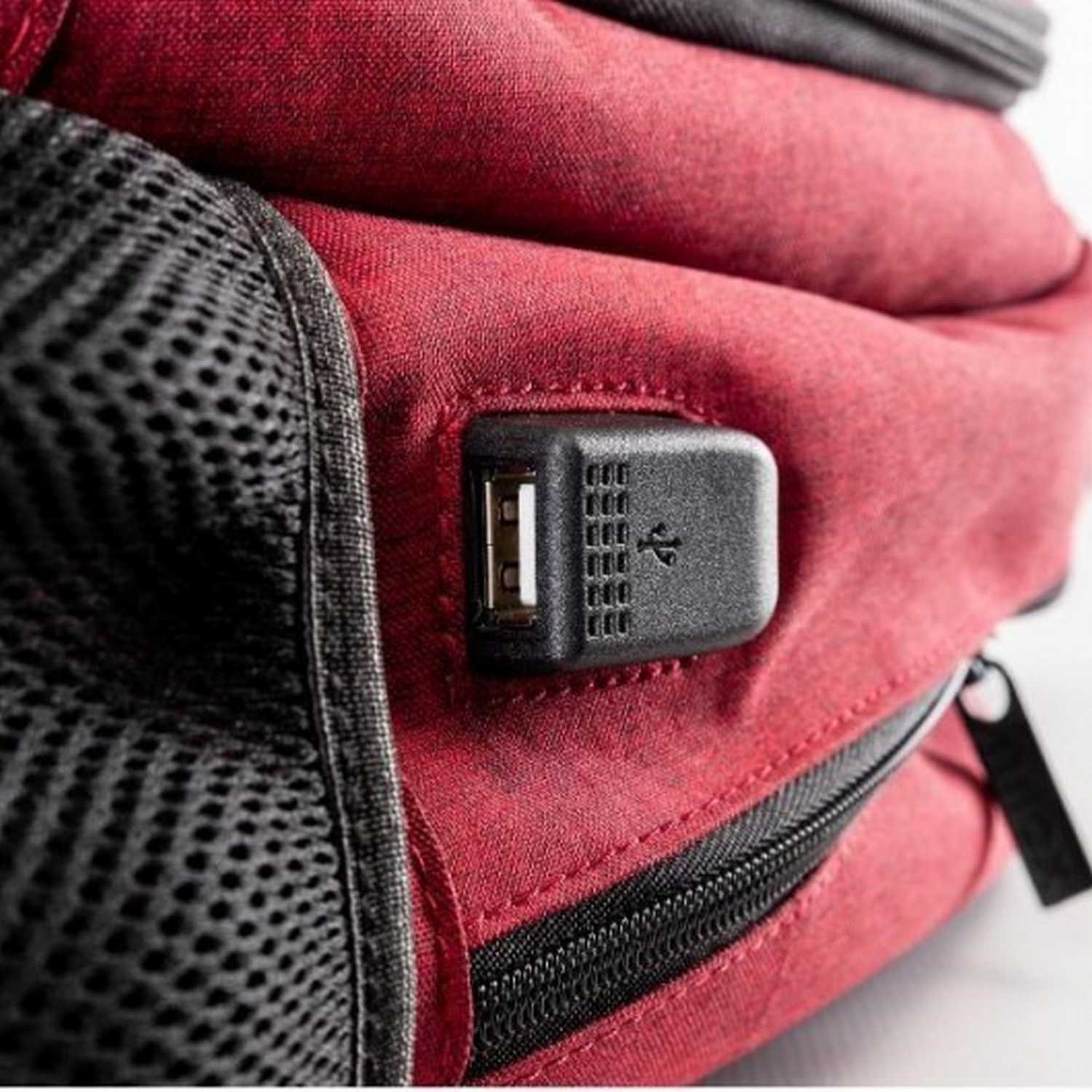 Detalle de mochila roja, con puerto USB y bolsillo de malla. Cremallera negra visible.