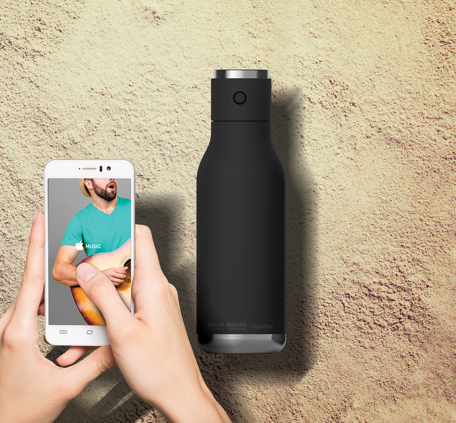 A person holding a phone with a music app open, with a black bottle next to it on sand.