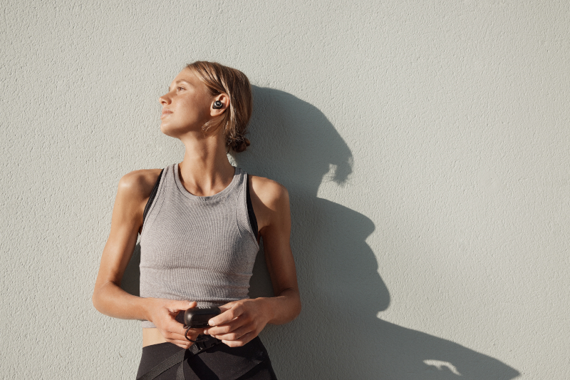 Blonde Frau in Sportkleidung steht an einer Wand und trägt Ohrhörer.