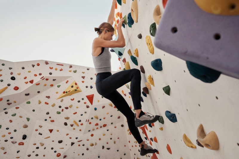 Frau klettert eine bunte Felswand. Sie trägt Leggings, ein Oberteil und Kopfhörer. Himmel im Hintergrund.