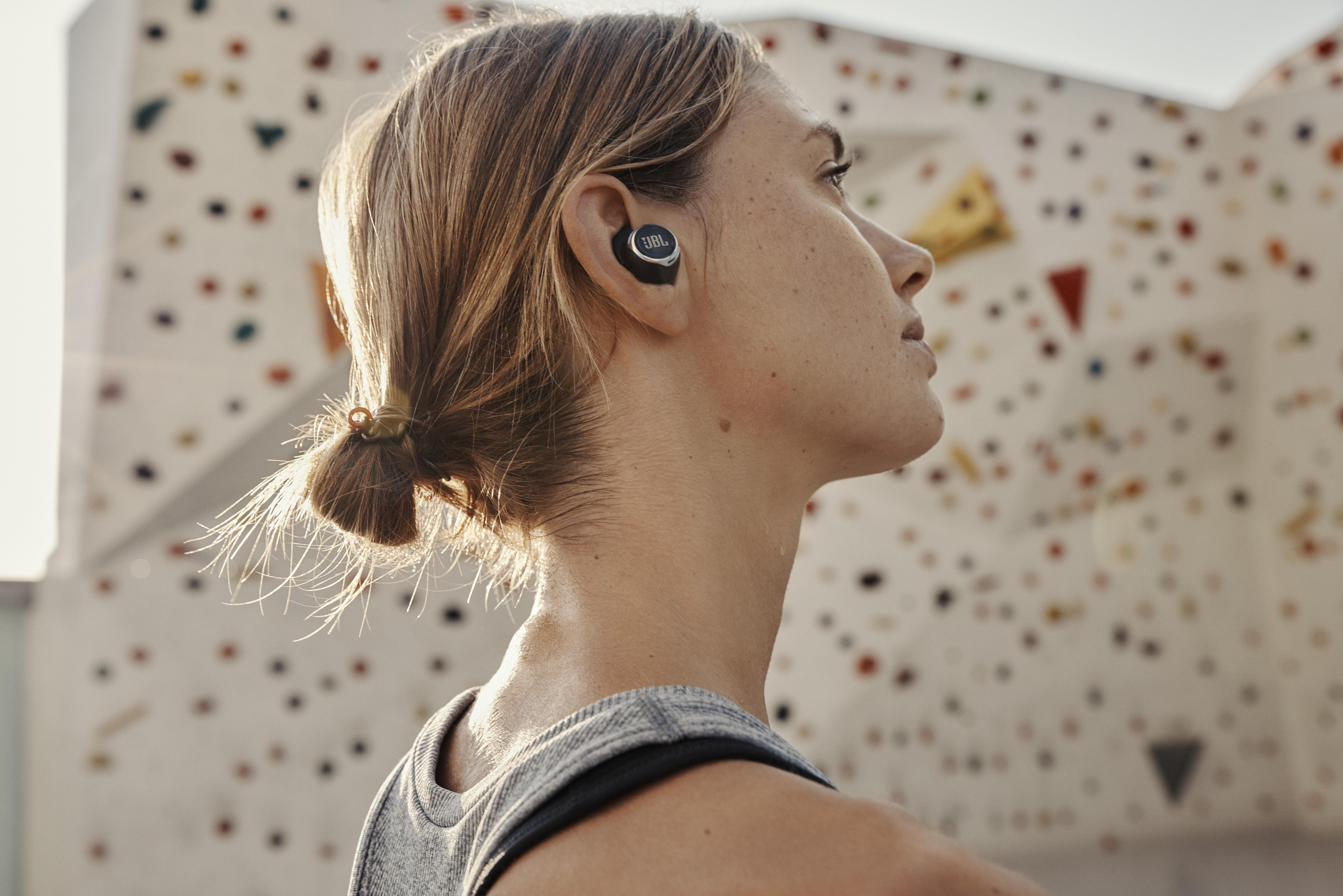 Frau mit JBL-Ohrhörern, nach oben schauend, Kletterwand im Hintergrund.