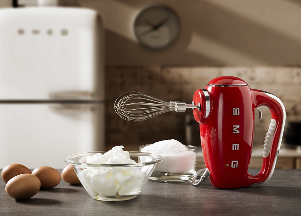 Frullatore a immersione SMEG rosso, ciotola con meringa, uova e zucchero su un tavolo.