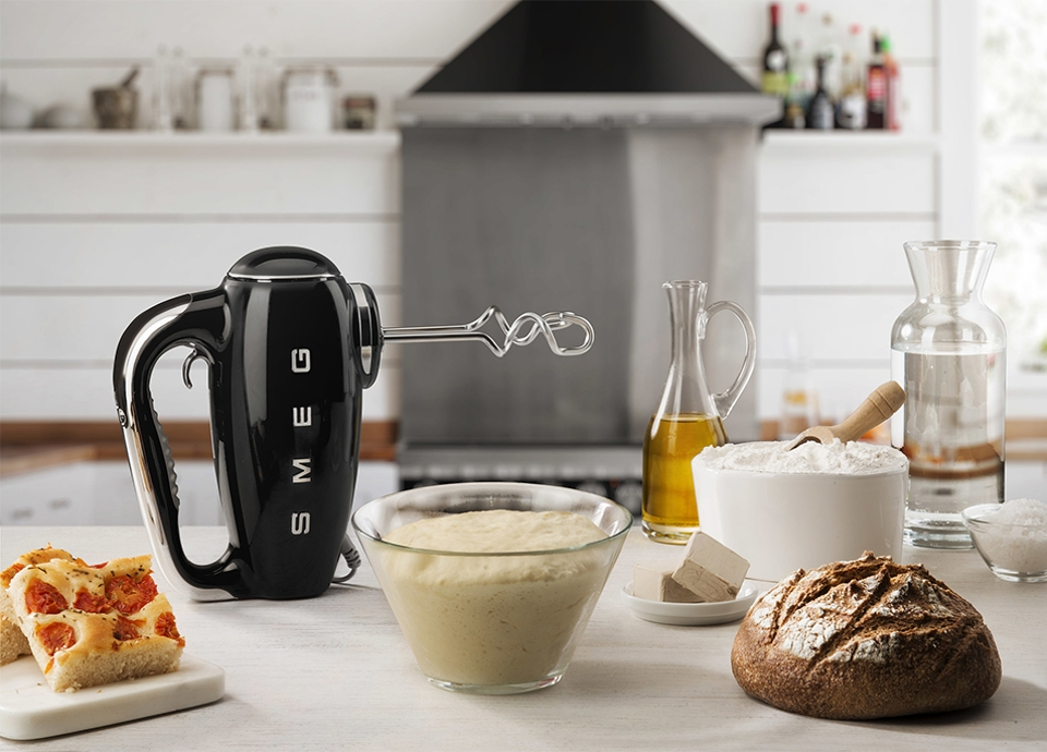 Un mixer a colonna SMEG nero con ingredienti su un piano di lavoro bianco in cucina.