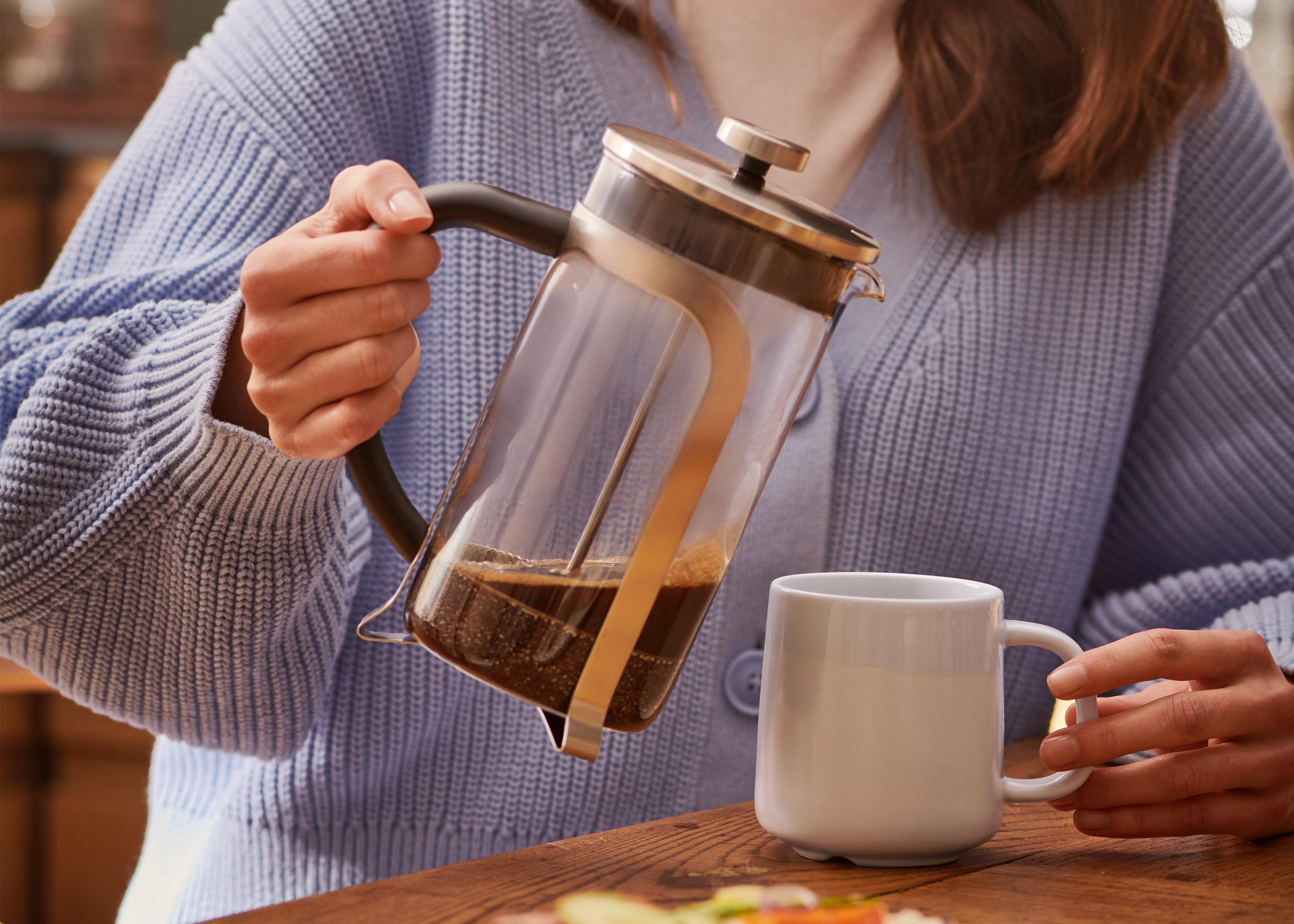 Eine Person gießt Kaffee aus einer French Press in eine weiße Tasse. Ein Holztisch und ein Fenster sind sichtbar.