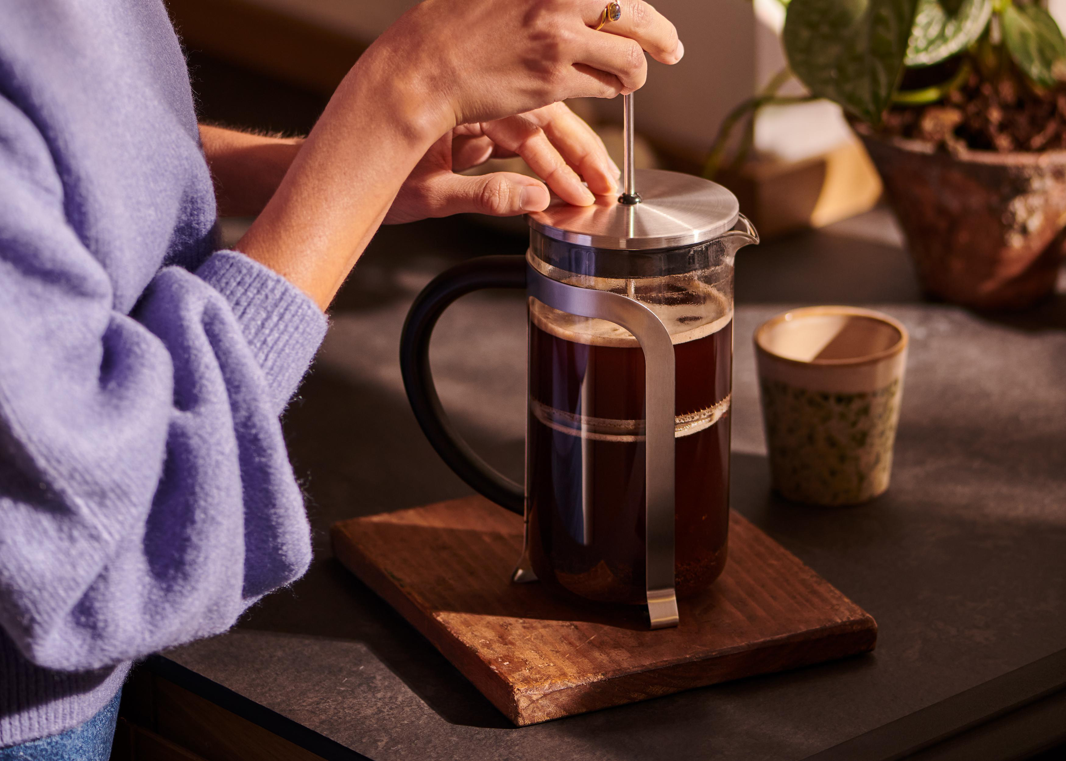 Eine Person drückt den Stempel einer French Press, die mit Kaffee gefüllt ist, auf einem Holztablett.