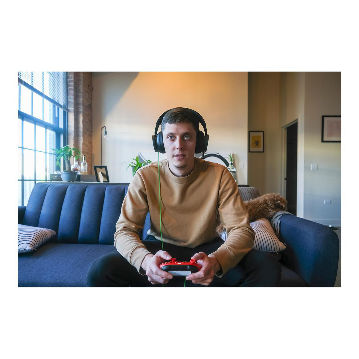 Ein Mann mit Kopfhörern hält einen roten Controller, während er auf einem blauen Sofa sitzt.