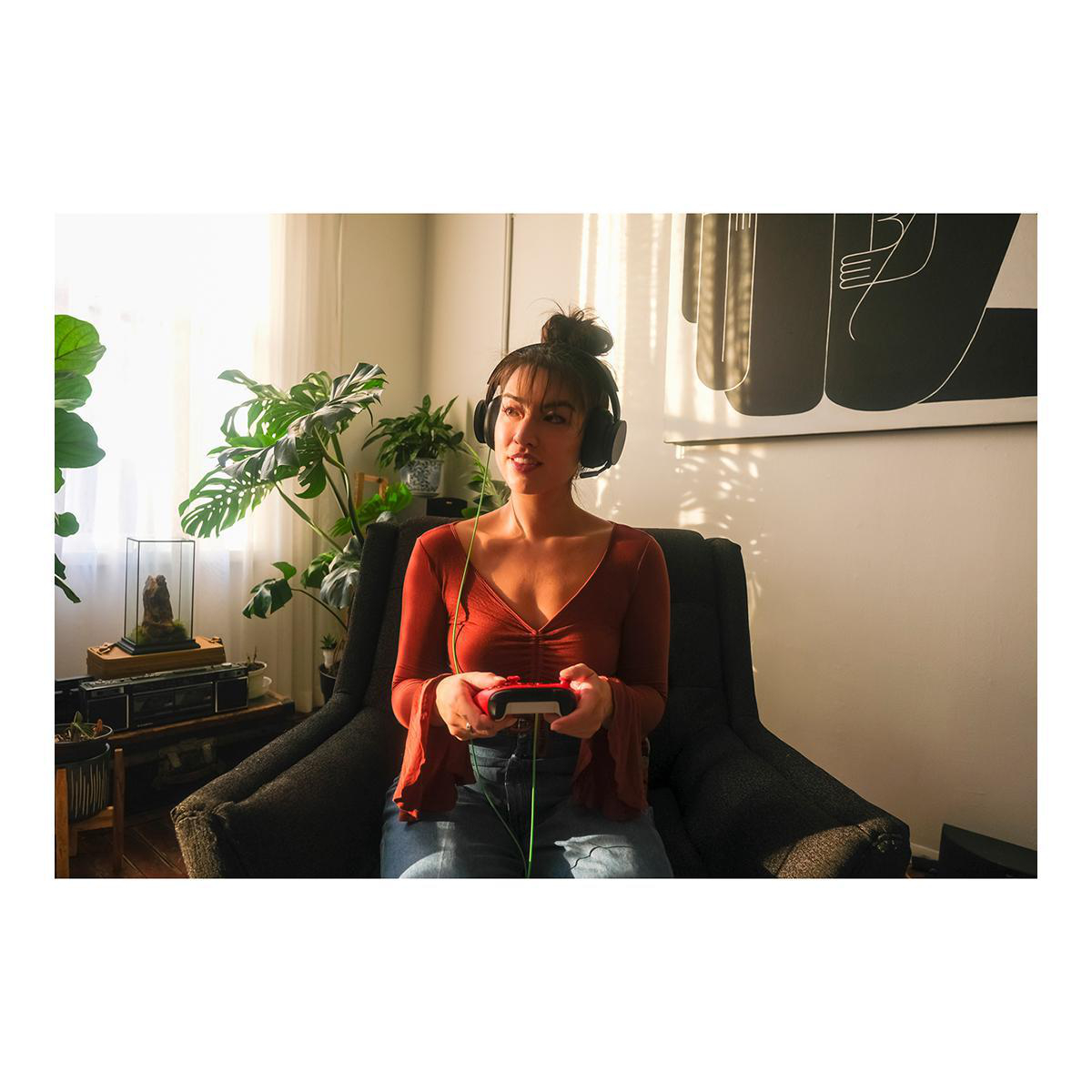 Frau in einem Raum, die mit einem Controller ein Videospiel spielt. Sie trägt Kopfhörer. Pflanzen sind im Hintergrund sichtbar.