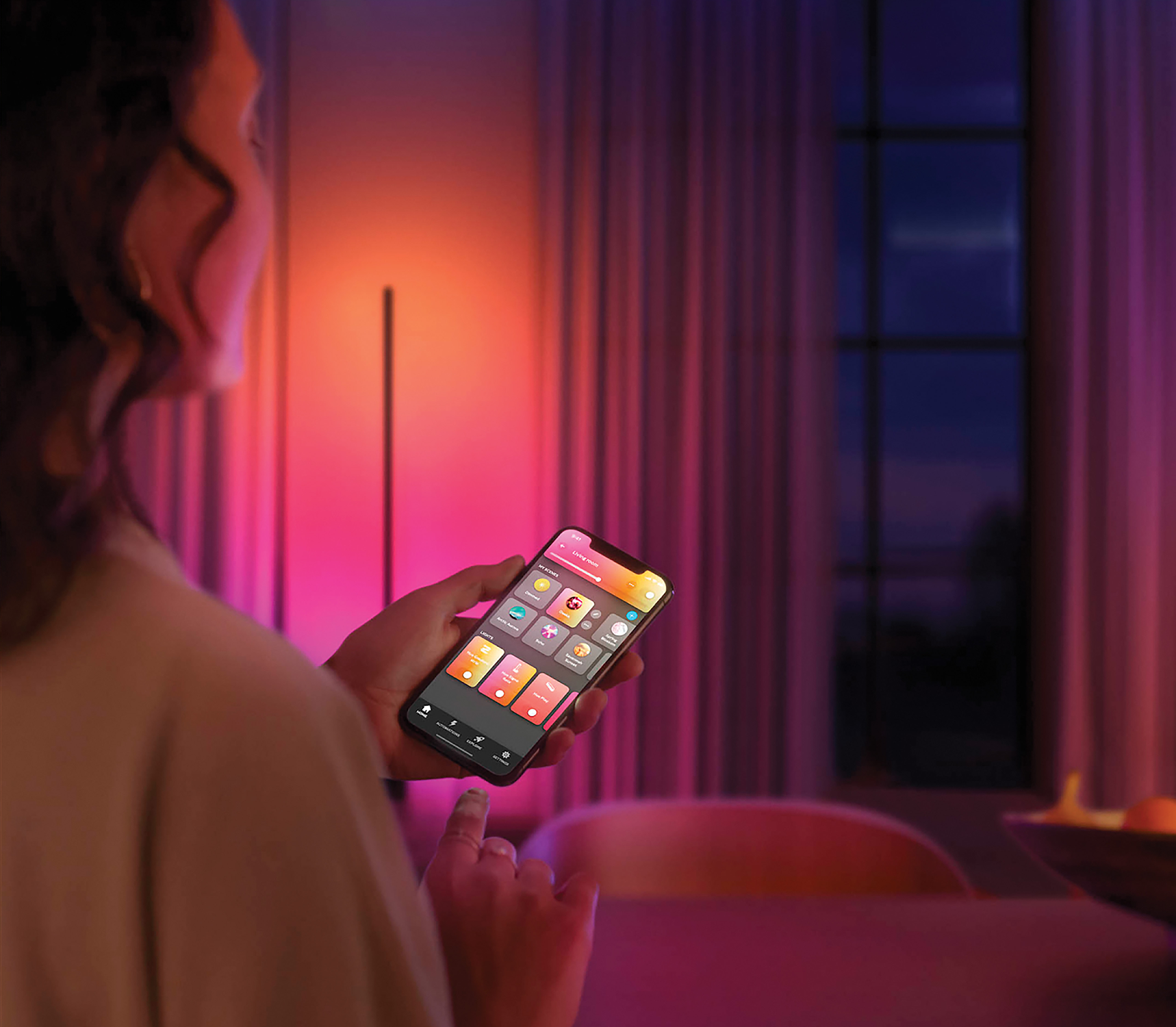 Een vrouw houdt een smartphone vast en communiceert met een smart home-app. De kamer is verlicht met gekleurde lichten.
