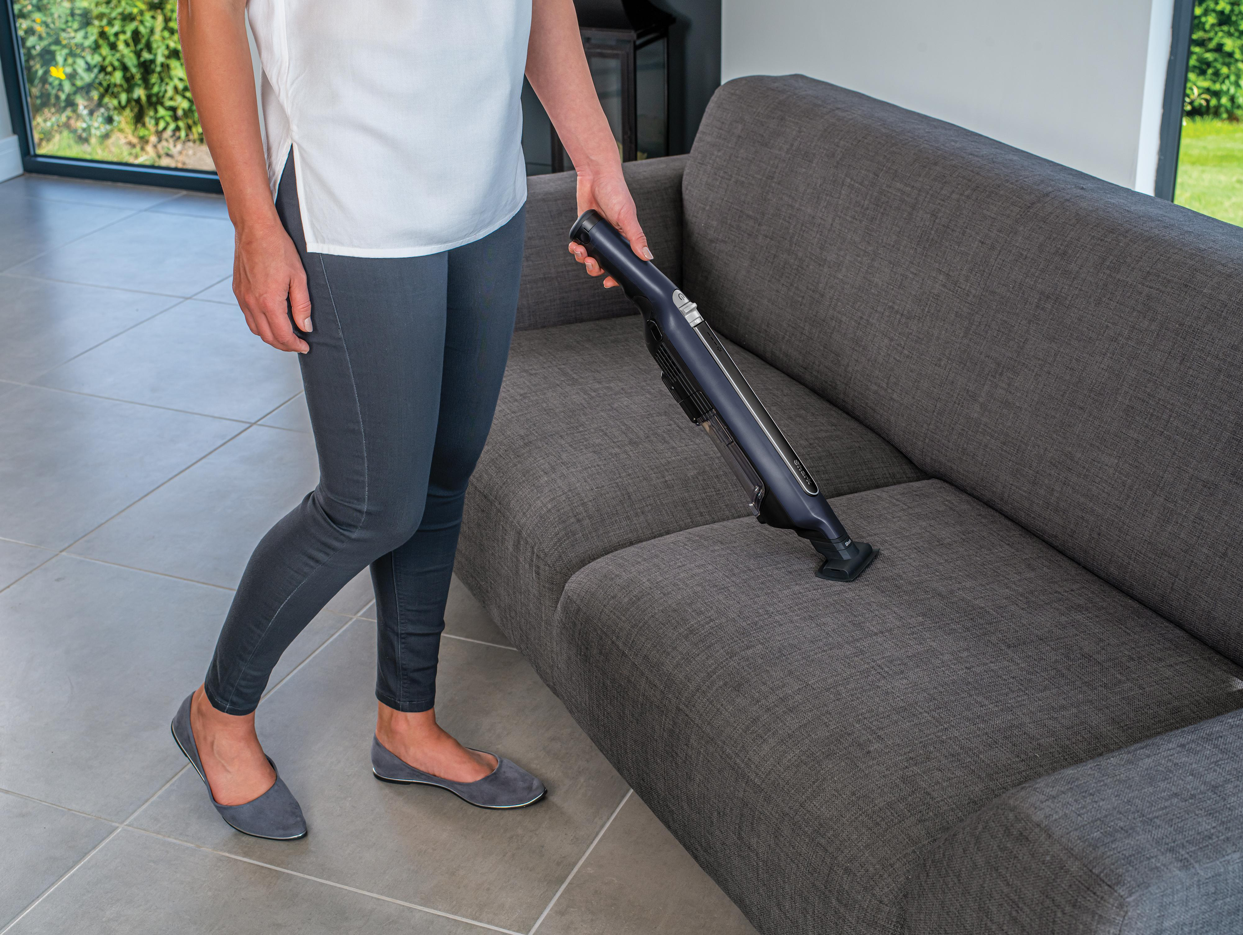 Eine Frau saugt mit einem Handstaubsauger ein graues Sofa ab.