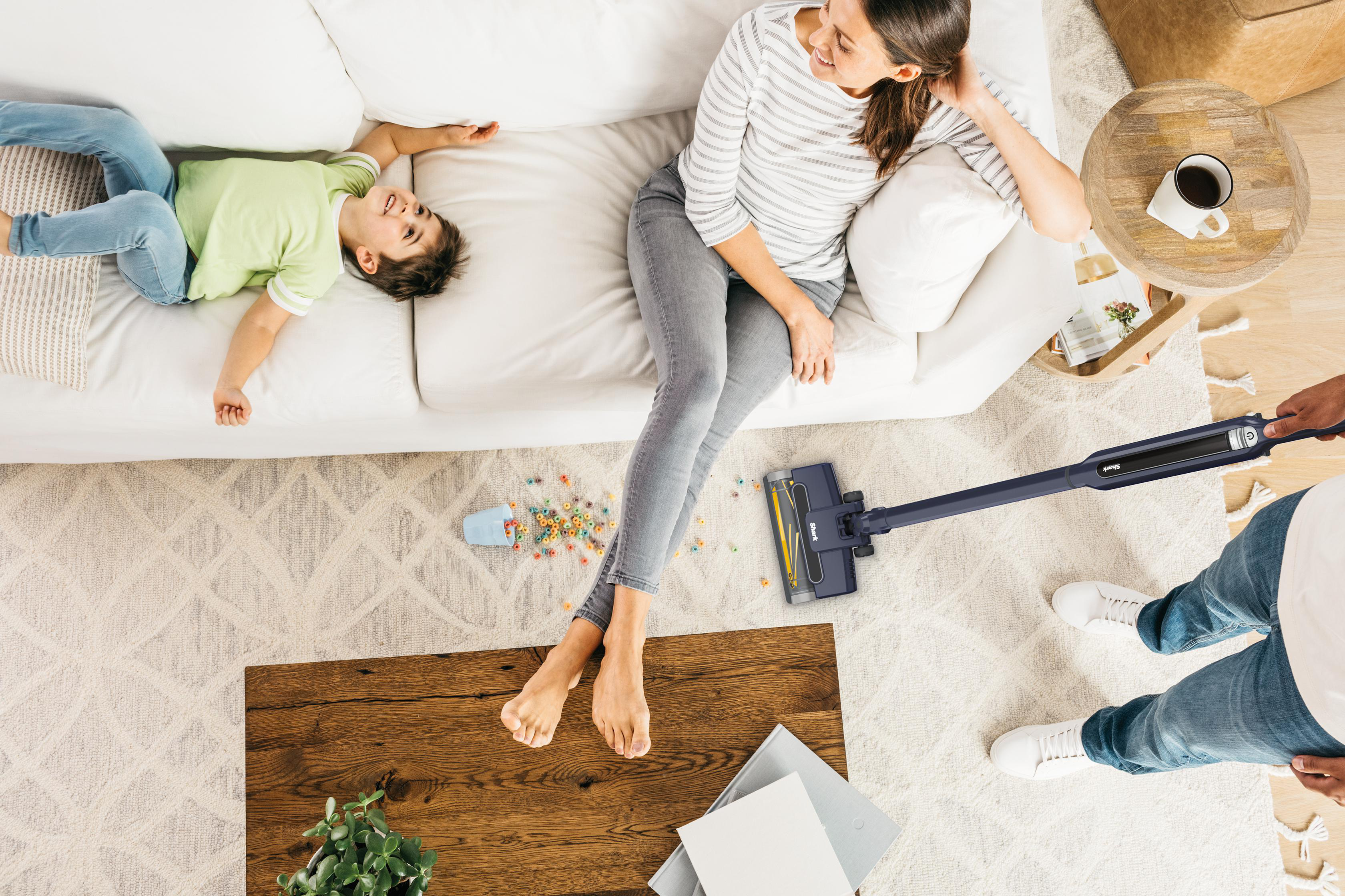 Eine Familie ist im Wohnzimmer. Mutter und Sohn sitzen auf der Couch. Vater saugt den Boden. Couchtisch und Teppich.