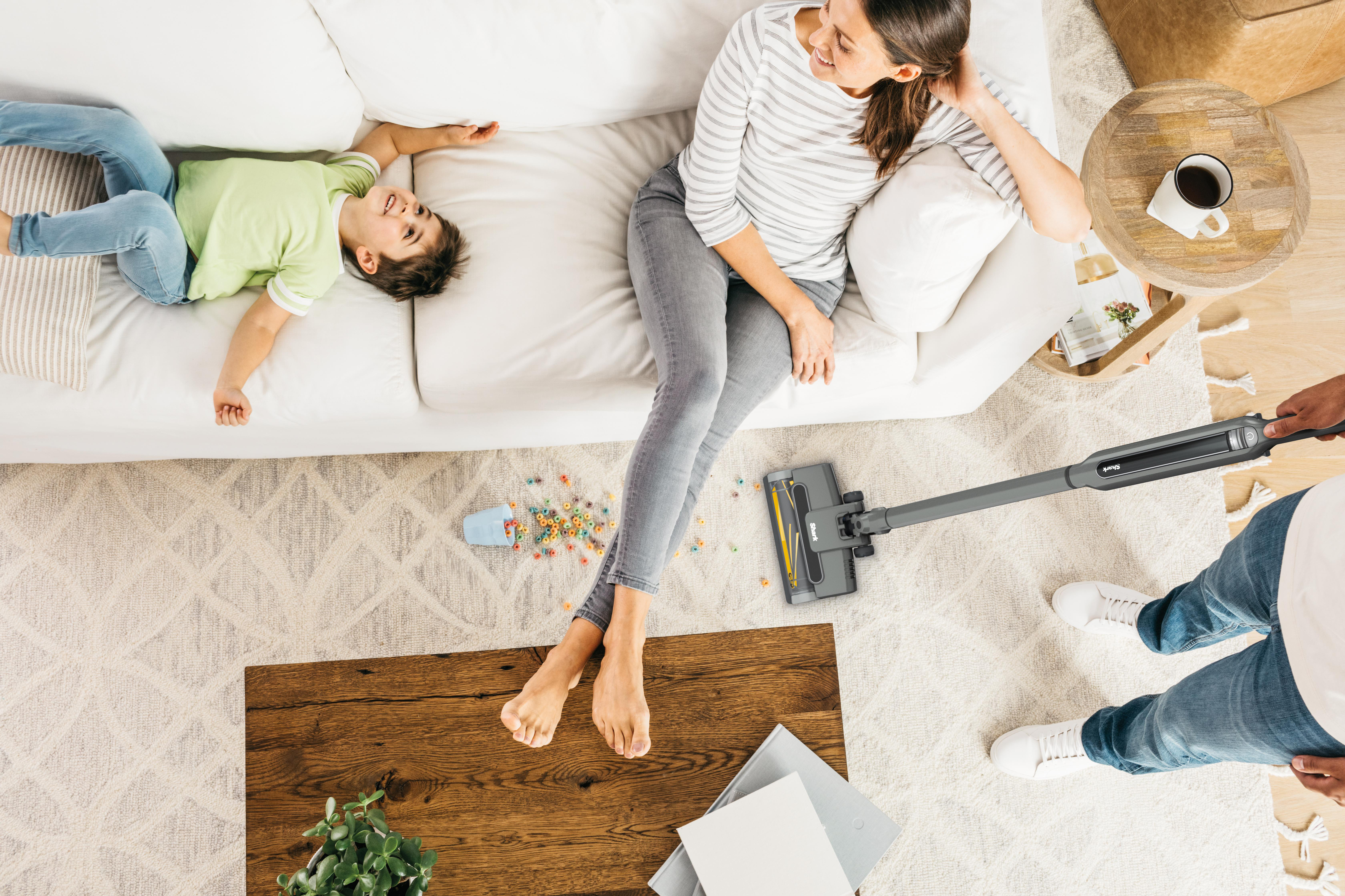 Eine Familie im Wohnzimmer. Eine Frau und ein Kind sitzen auf dem Sofa. Ein Mann saugt den Boden.