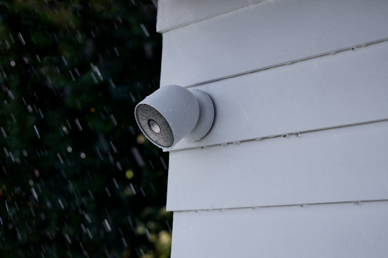 Eine Überwachungskamera an einem weiß getäfelten Haus, Regen fällt darauf.