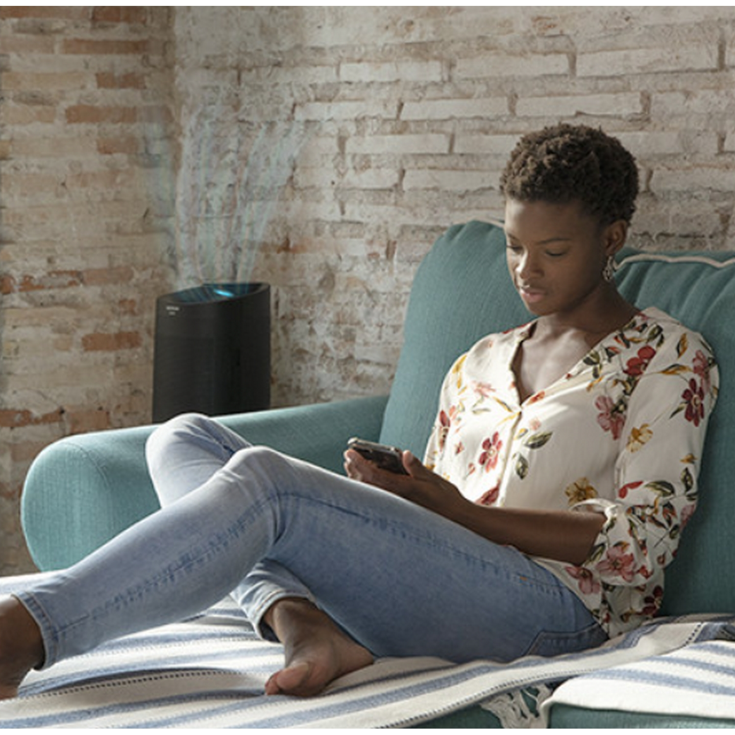 Mujer en un sofá, usando un teléfono. Un purificador de aire negro está a la izquierda.