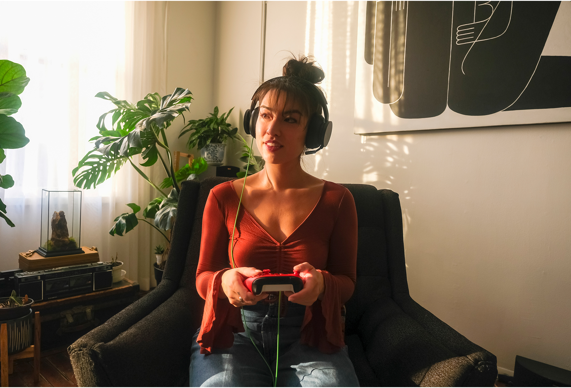 Femme en chemise rouge avec casque jouant à un jeu. Plantes et art en arrière-plan. La lumière du soleil traverse une fenêtre.