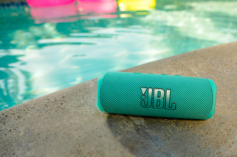 Ein türkisfarbener JBL-Lautsprecher liegt am Poolrand. Pinke und gelbe Schwimmer im Hintergrund, Wasser.