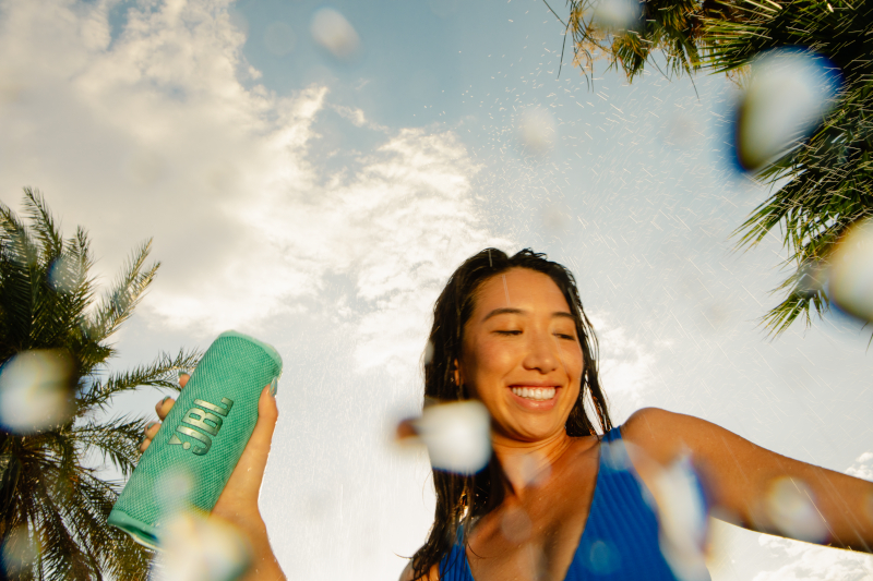 Frau hält grünen JBL-Lautsprecher, Wasser spritzt in der Luft, blauer Himmel im Hintergrund.