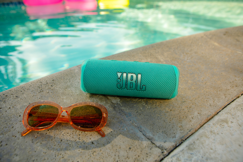Türkisgrüner JBL-Lautsprecher und orangefarbene Sonnenbrille auf Beton neben Pool.