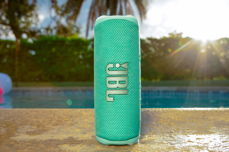 Ein türkisfarbener JBL-Lautsprecher steht auf einem Sims in der Nähe eines Pools. Die Sonne scheint hell im Hintergrund.