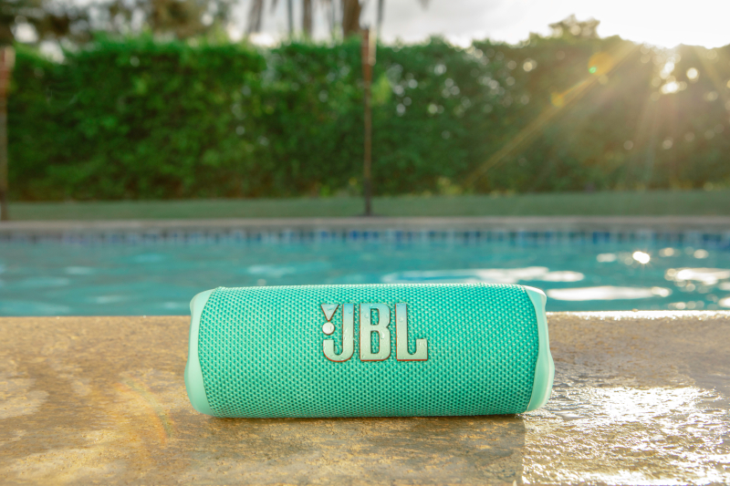 Ein türkisblauer JBL-Lautsprecher auf einer Steinoberfläche mit Pool und Sonnenlicht im Hintergrund.