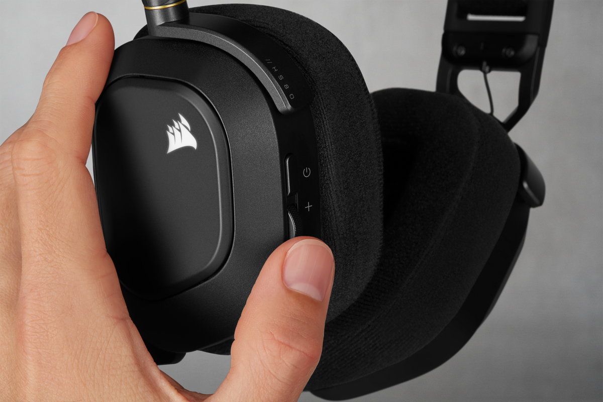 Hand hält schwarzes Corsair-Headset. Lautstärketasten sichtbar. Grauer Hintergrund.