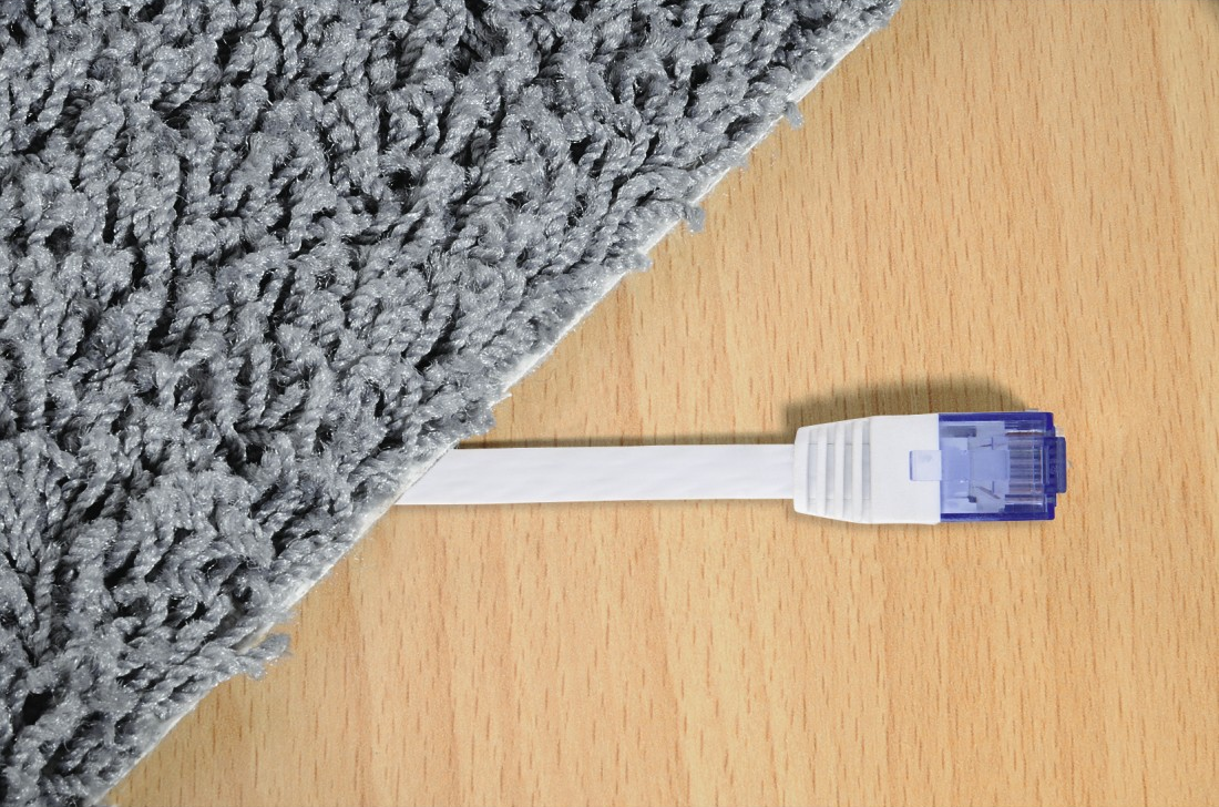 Un cavo di rete bianco con connettore blu si estende da sotto un tappeto grigio su una superficie di legno.