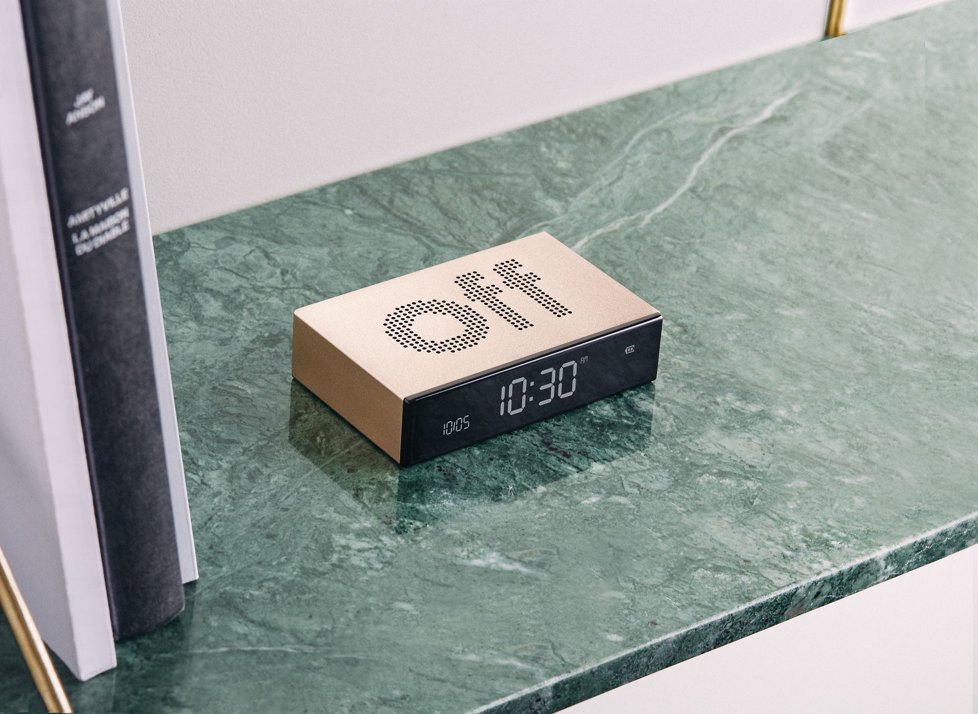 Sveglia dorata con 'off' in alto, su un ripiano di marmo verde con libri a sinistra.