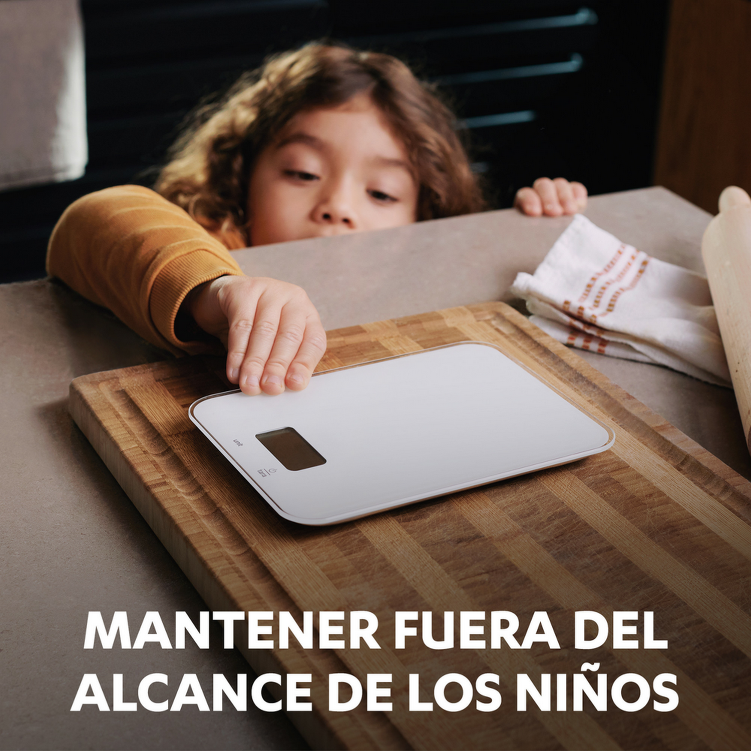 Un niño alcanza una báscula de cocina digital blanca sobre una tabla de cortar de madera. Texto: Mantener fuera del alcance de los niños.