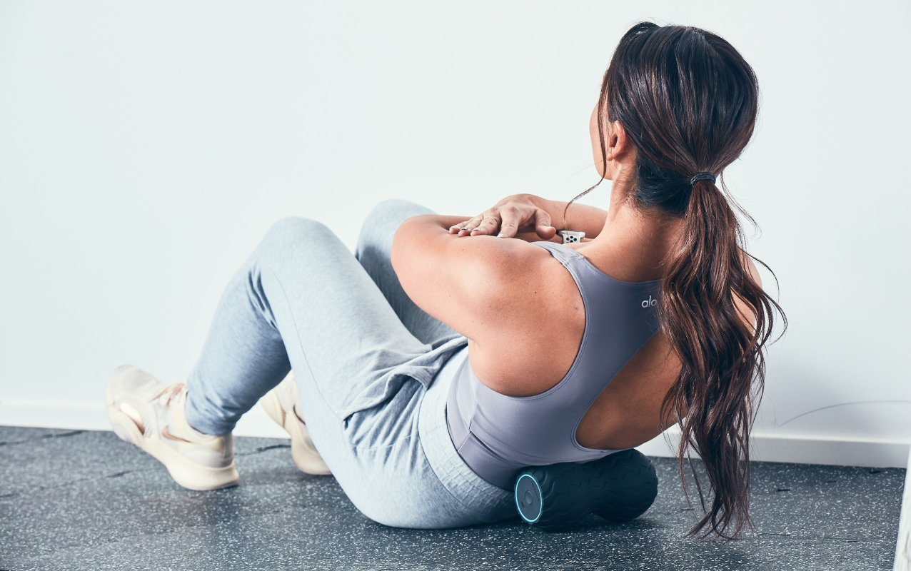 Frau in grauer Trainingskleidung, die Sit-ups auf einer schwarzen Schaumstoffrolle macht, in der Nähe einer weißen Wand.