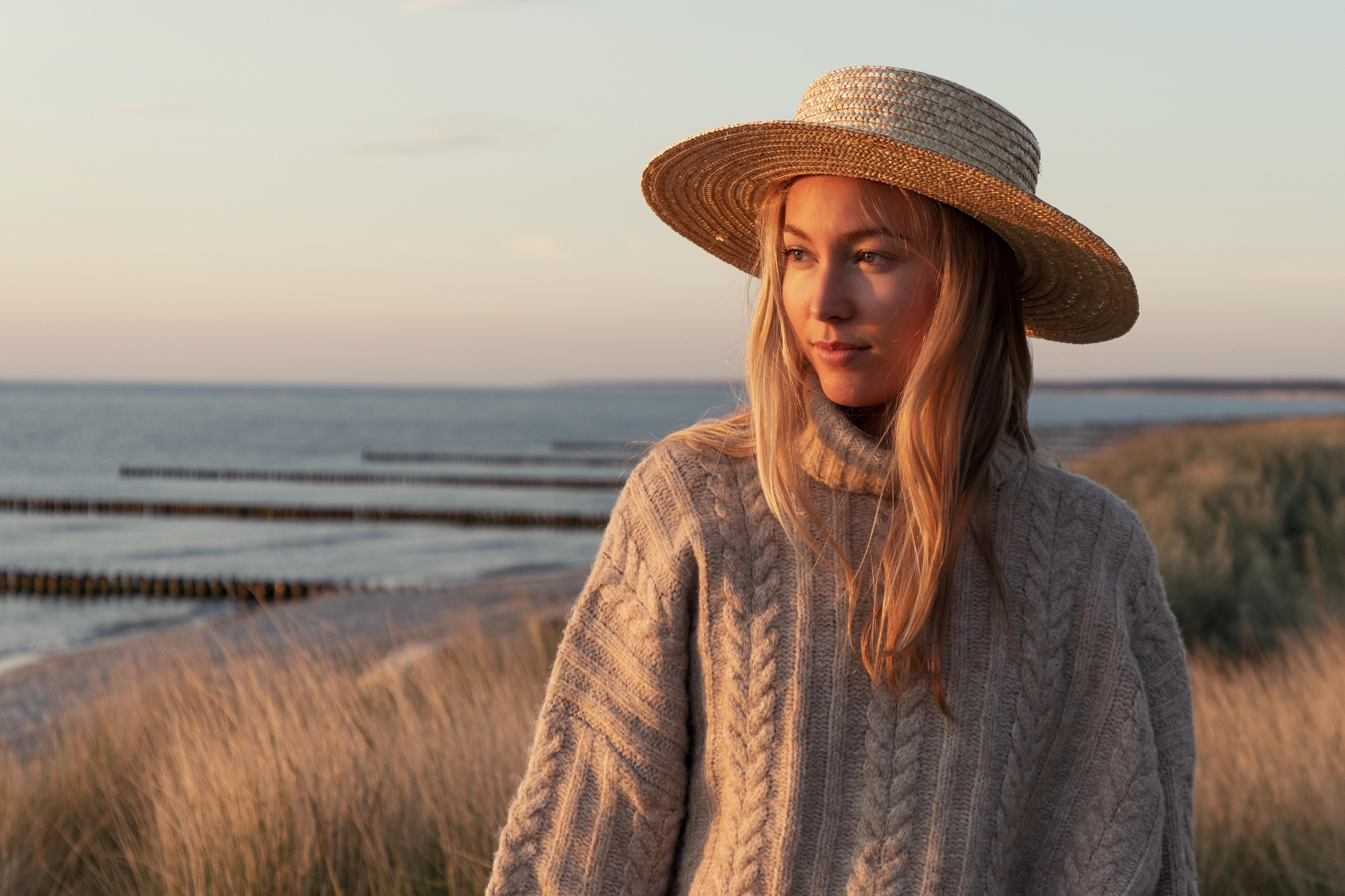 Eine Frau mit Hut und Pullover steht auf einem Hügel und blickt aufs Meer.