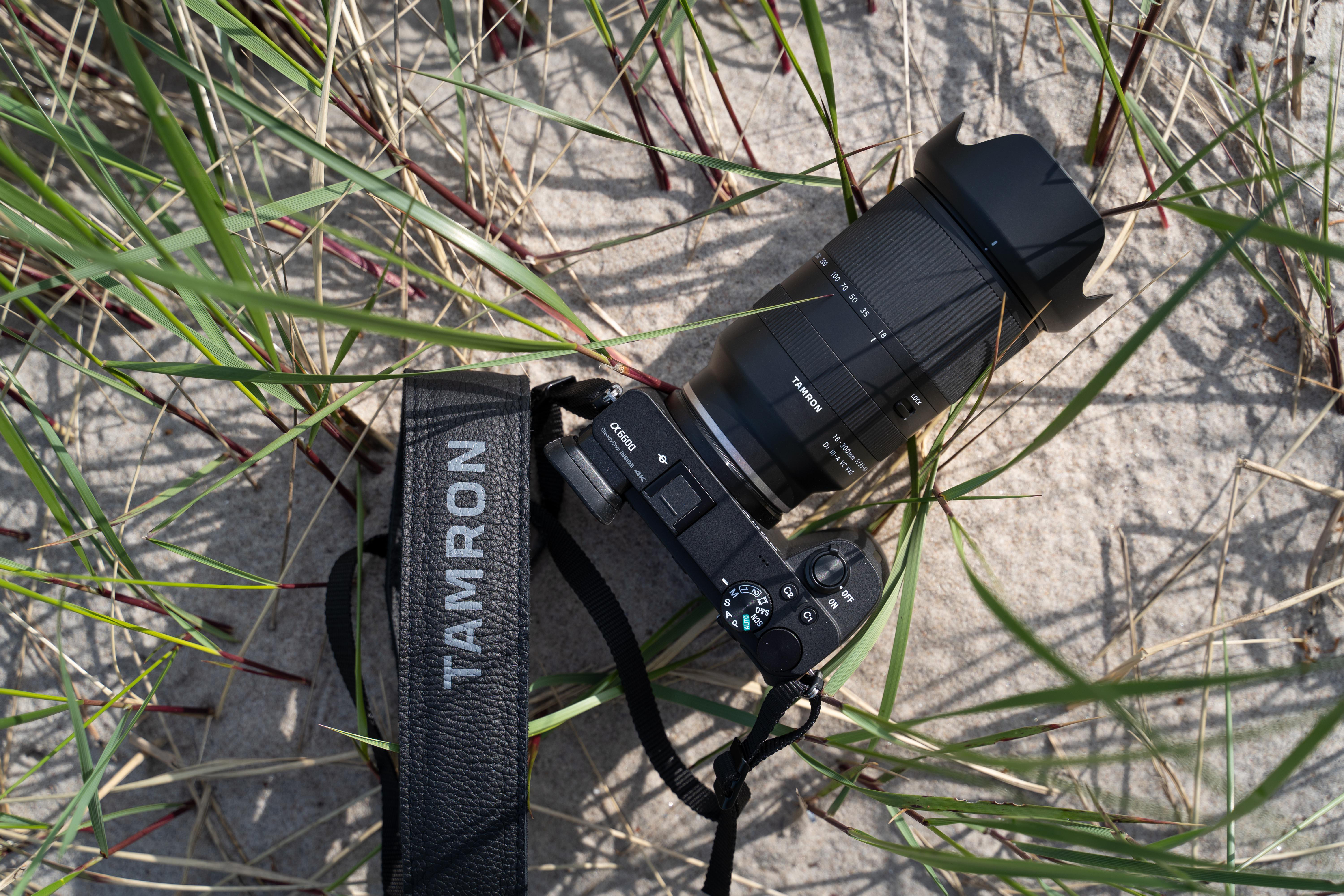 Kamera auf Sand mit grünem Gras. Schwarze Kamera mit Tamron-Objektiv und -Gurt.