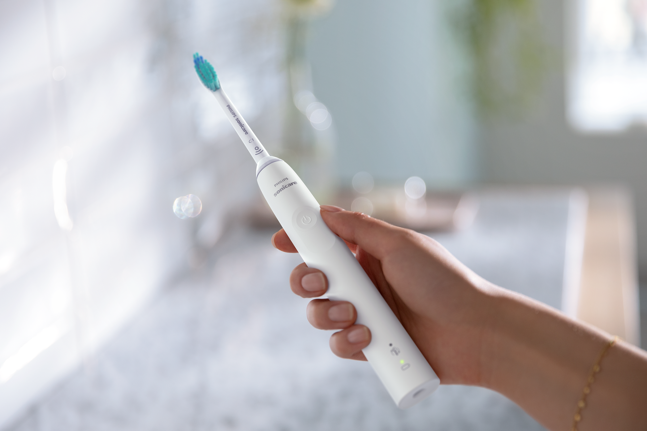 Hand holding a white Philips Sonicare toothbrush with a blue-green brush head, bathroom background.