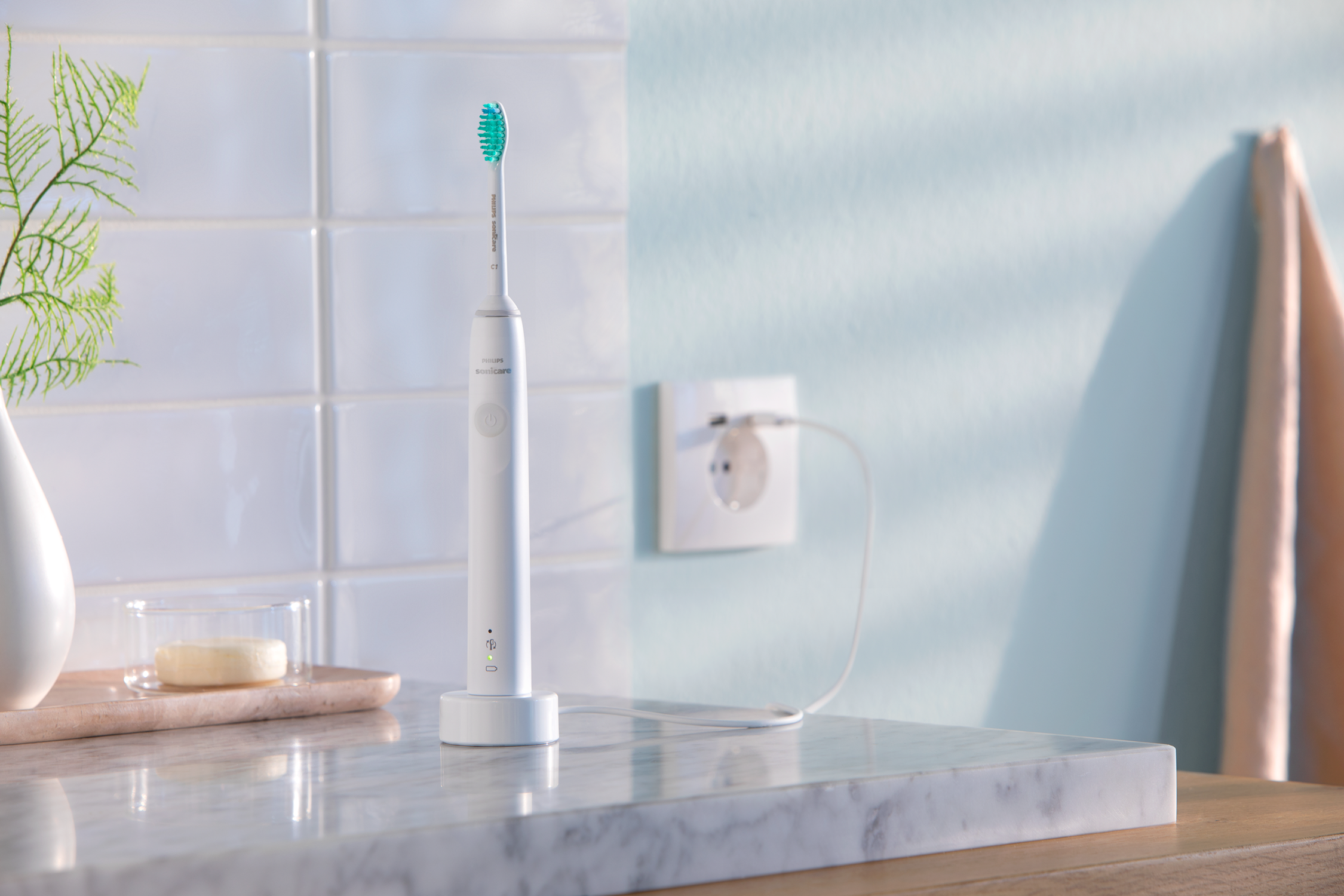 White electric toothbrush on a charging base, green bristles. Tiled wall and a plug.