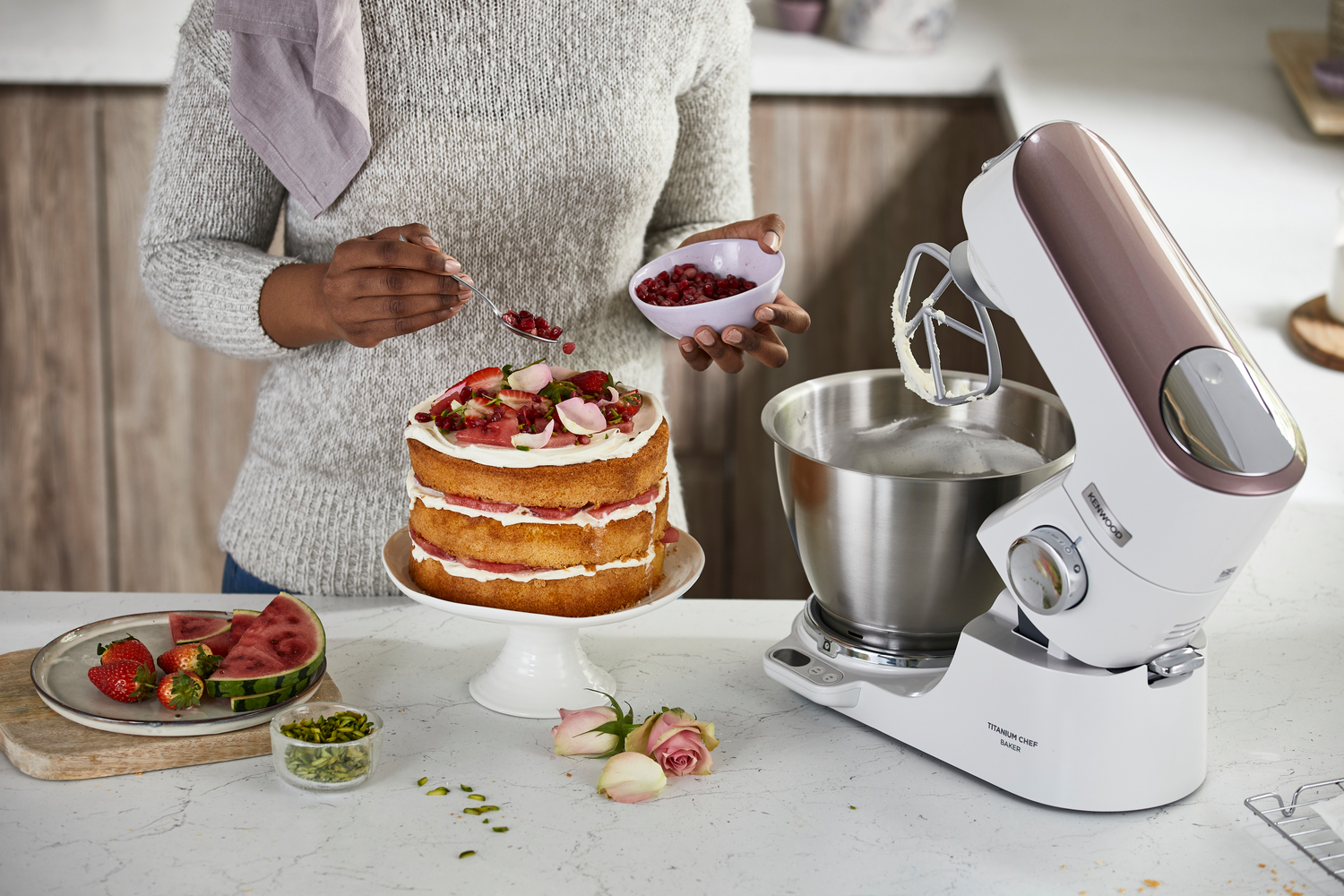 Eine Person dekoriert einen Kuchen mit Erdbeeren und Pistazien. Ein Mixer steht auf der Theke.