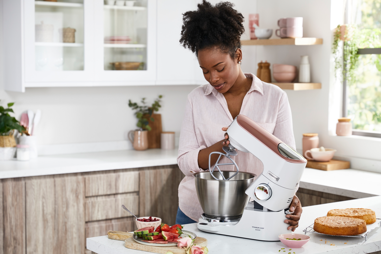 Eine Frau in einer Küche benutzt einen weißen und kupferfarbenen Küchenmixer, mit Kuchen und Obst in der Nähe.