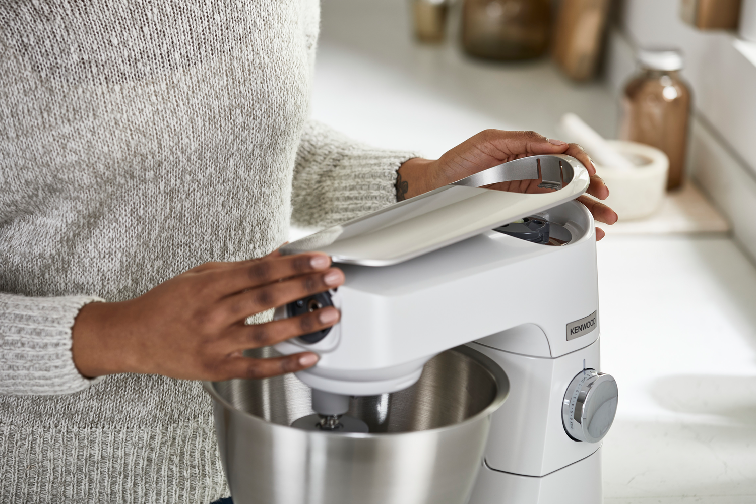 Hände einer Person öffnen den Deckel einer weißen Kenwood Küchenmaschine, in einer Küche stehend.
